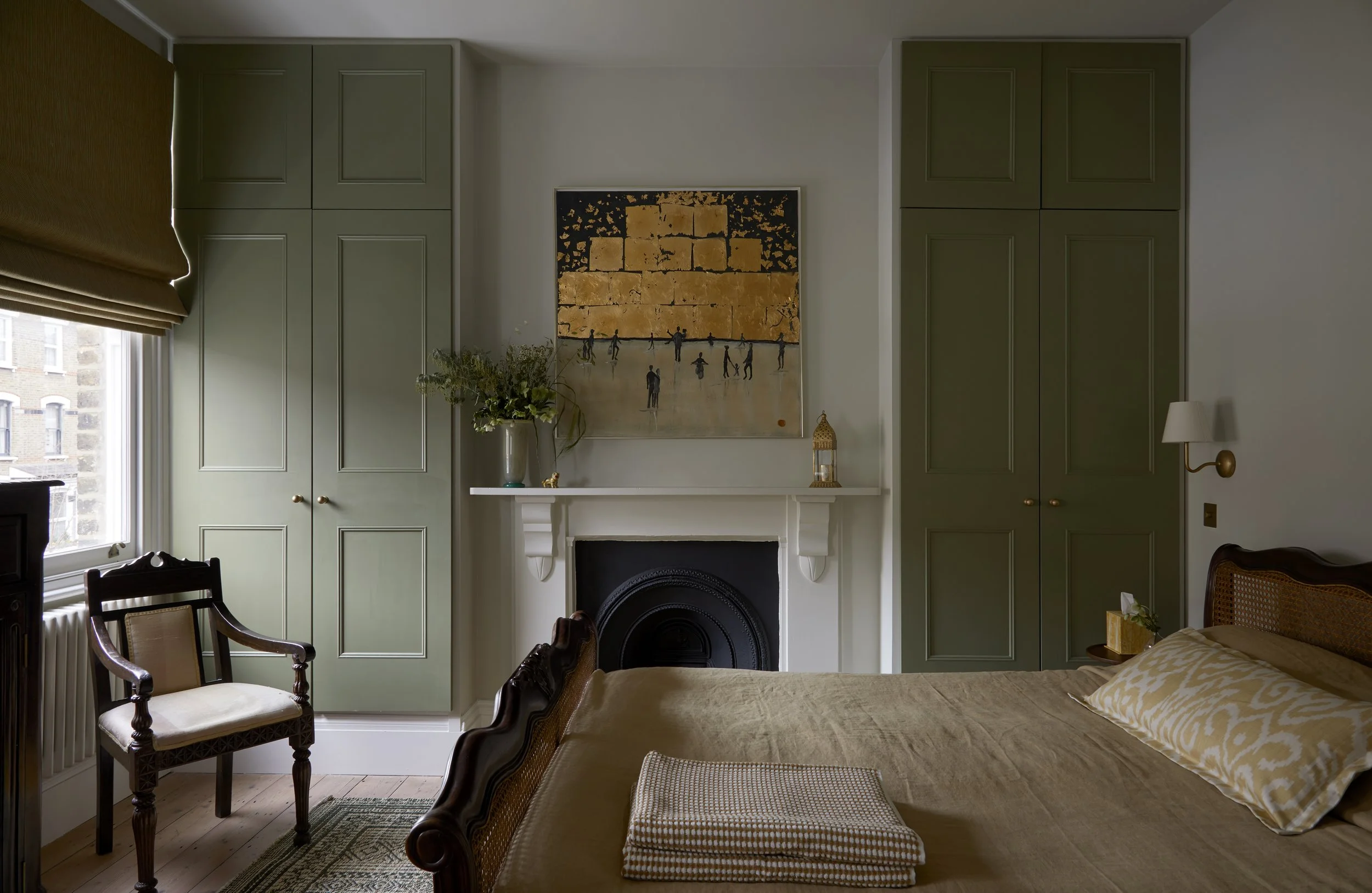 A cozy bedroom featuring a white fireplace with decorative shelving, leafy green vase, and artwork above, flanked by built-in green wardrobes, a window with brown Roman shade, a vintage armchair, a bed with  bedding and patterned pillow, a nightstand