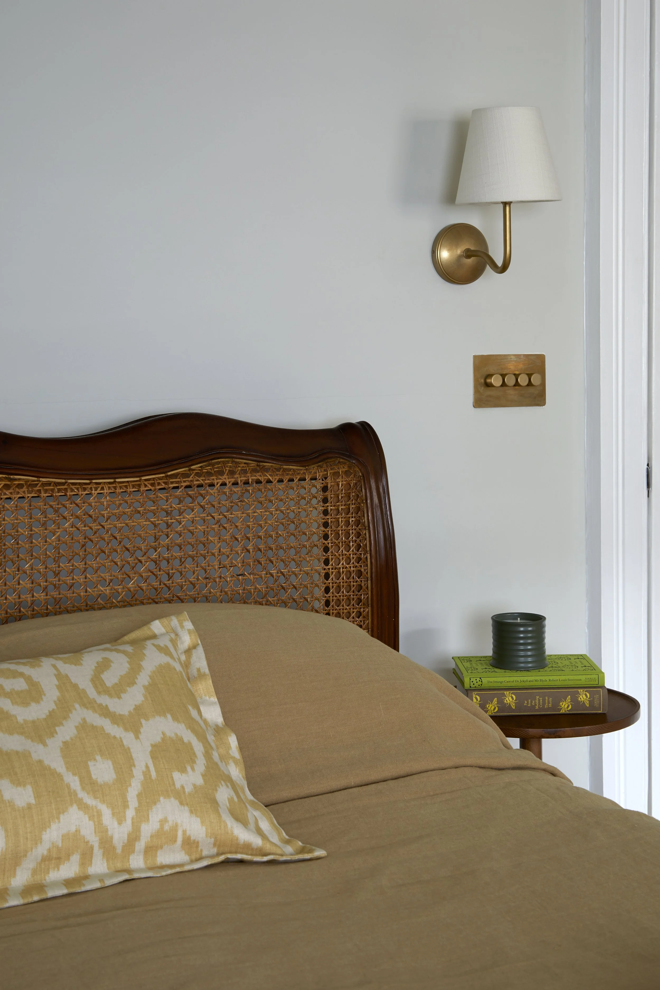 Close-up of a bedroom corner featuring a wooden headboard with woven cane detail, a bedspread, and a yellow patterned pillow. A small wooden side table holds green and yellow books and a black candle holder. Above, a wall-mounted brass and fabric wal