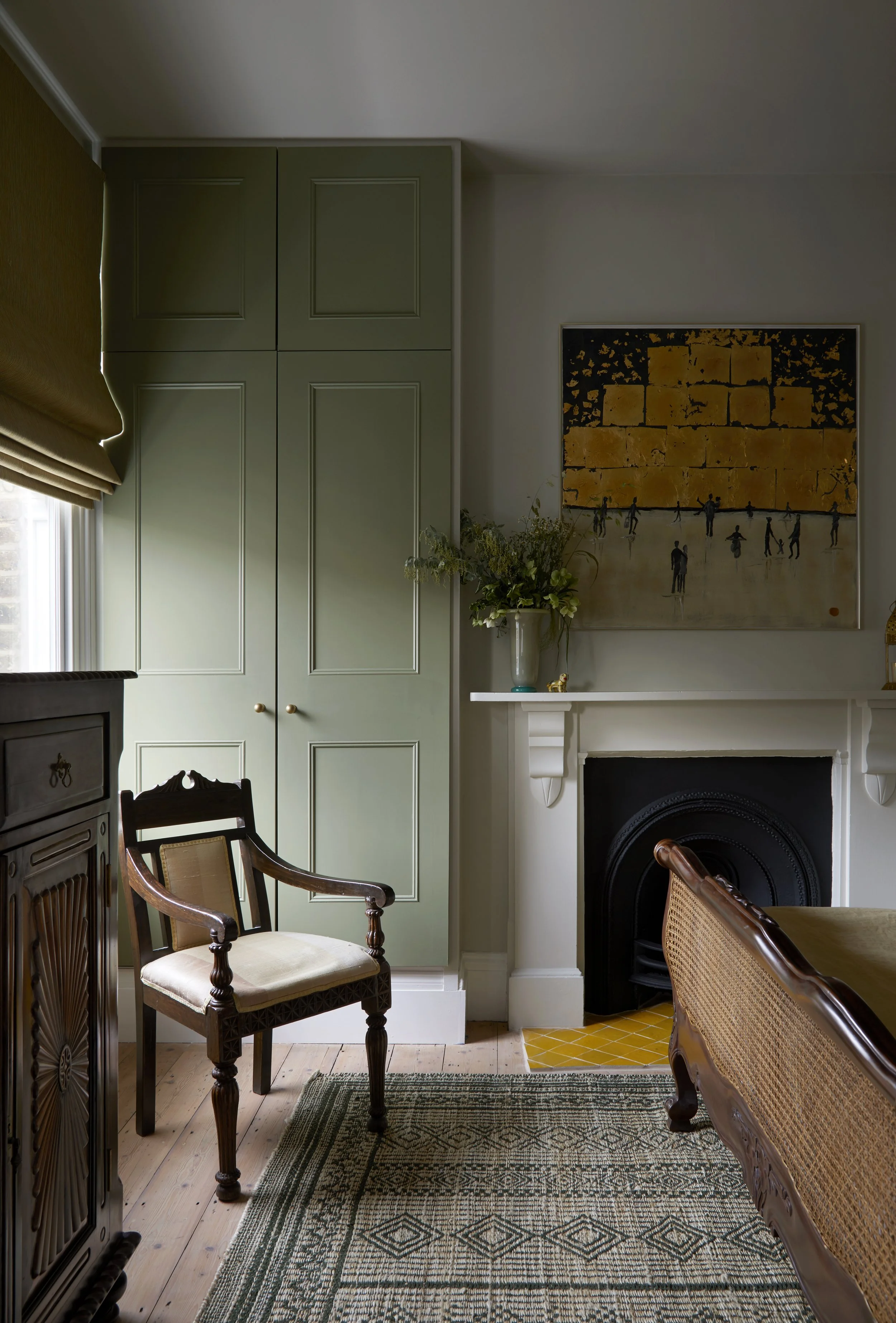 Interior of a living room with green built-in cabinets, a decorative fireplace, an armchair with a beige cushion, a wooden dining table, a patterned rug, a window with beige curtains, and art on the wall.