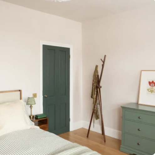 A bedroom with a green door, a bed with white and green striped bedding, a small wooden nightstand with a green lamp, a coat rack with dressing gown, and a green dresser with a framed flower artwork on top.