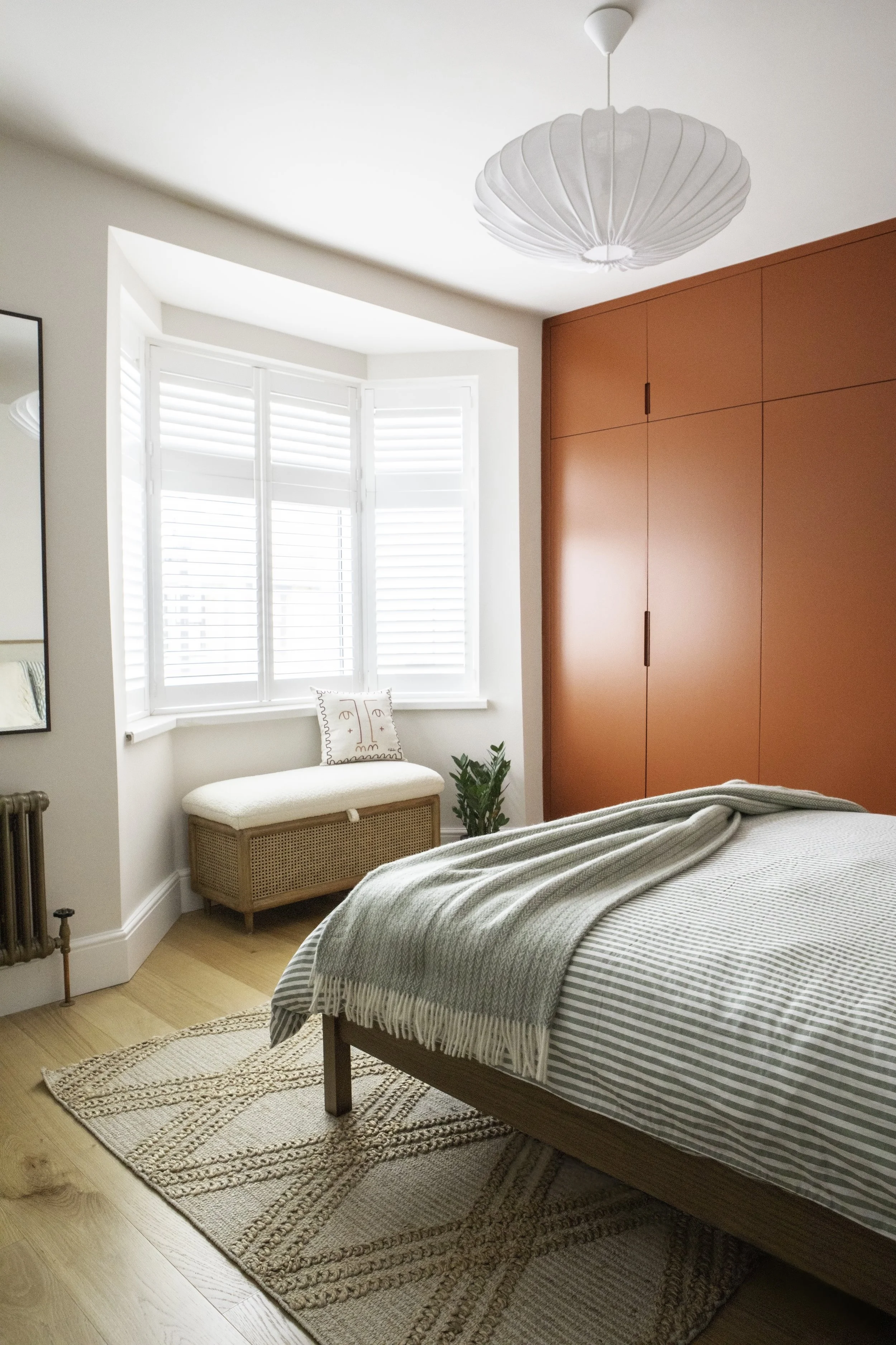 A bedroom with a window, a bench with a pillow, and a bed with a throw blanket. There is a  wardrobe in Farrow and Ball, a ceiling lamp, a mirror, and a plant.