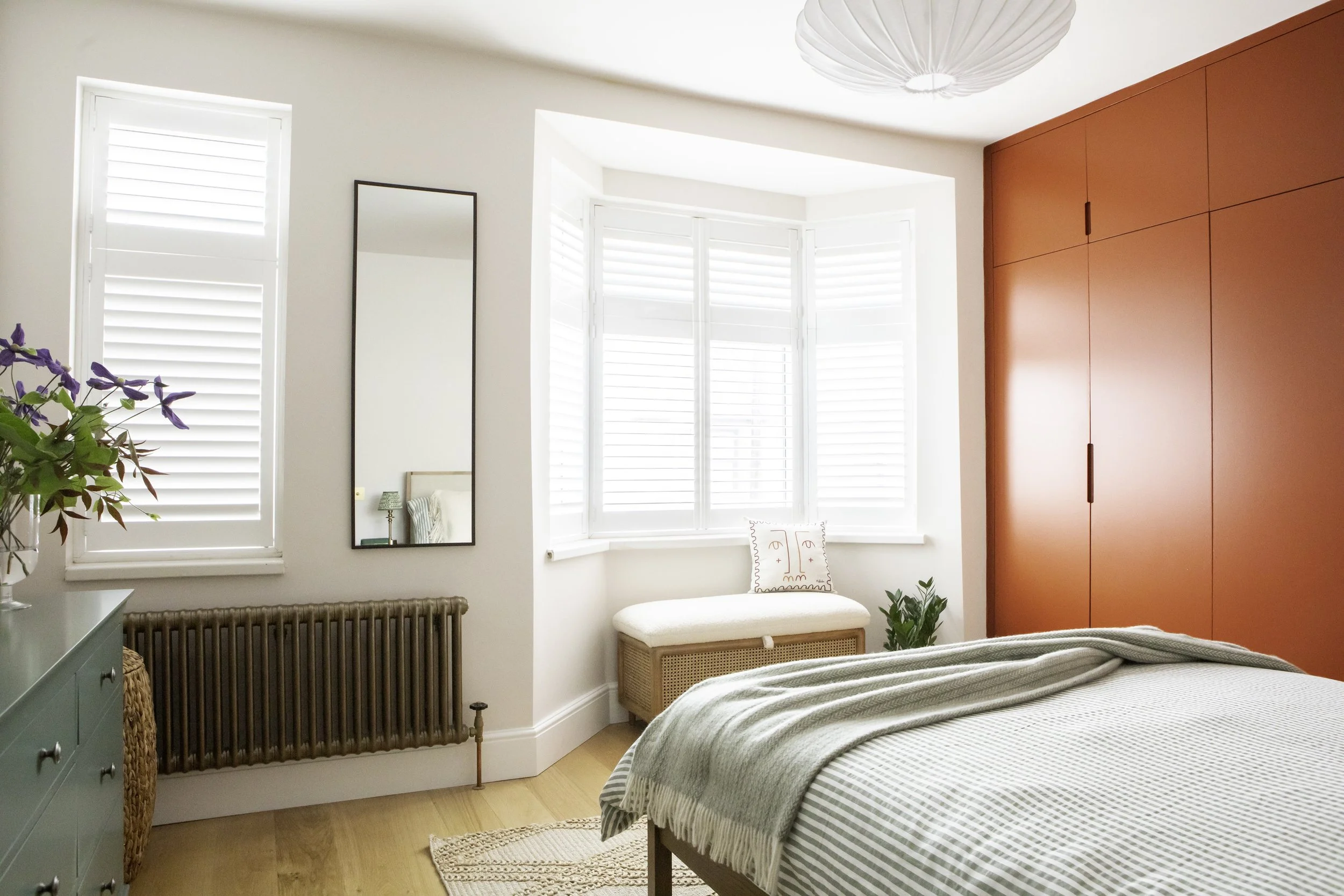 Bright bedroom with bay window, white shutters, and a red wardrobe. Bed with green and white striped bedding, a cream-colored blanket, and decorative pillow. Green dresser with purple flowers, mirror, and a small bench with a cushion near the window.