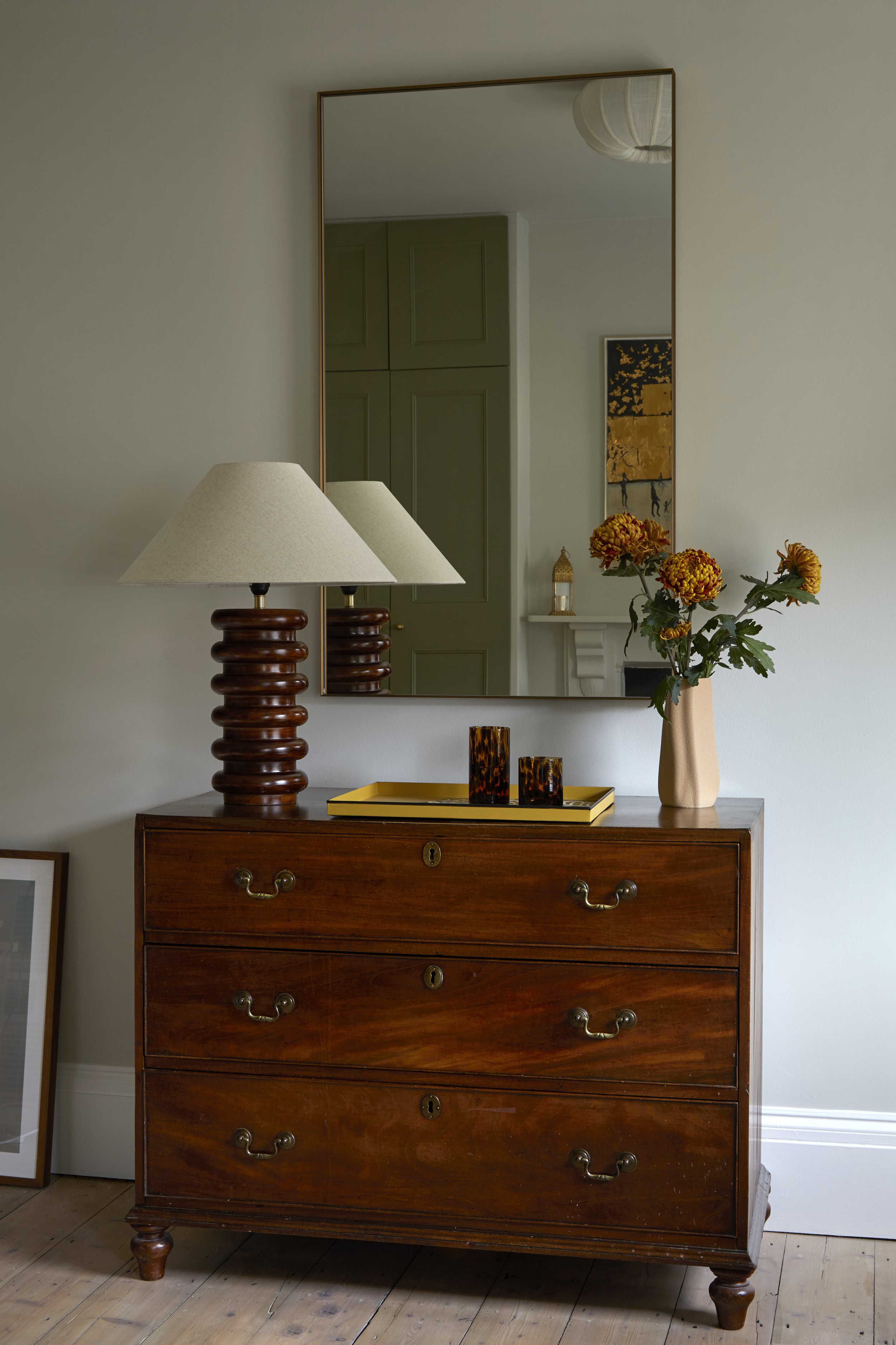 A wooden dresser with brass handles, a table lamp with a ribbed wooden base, a yellow tray with decor items, and a vase with flowers, a mirror above, and a framed picture leaning on the wall.