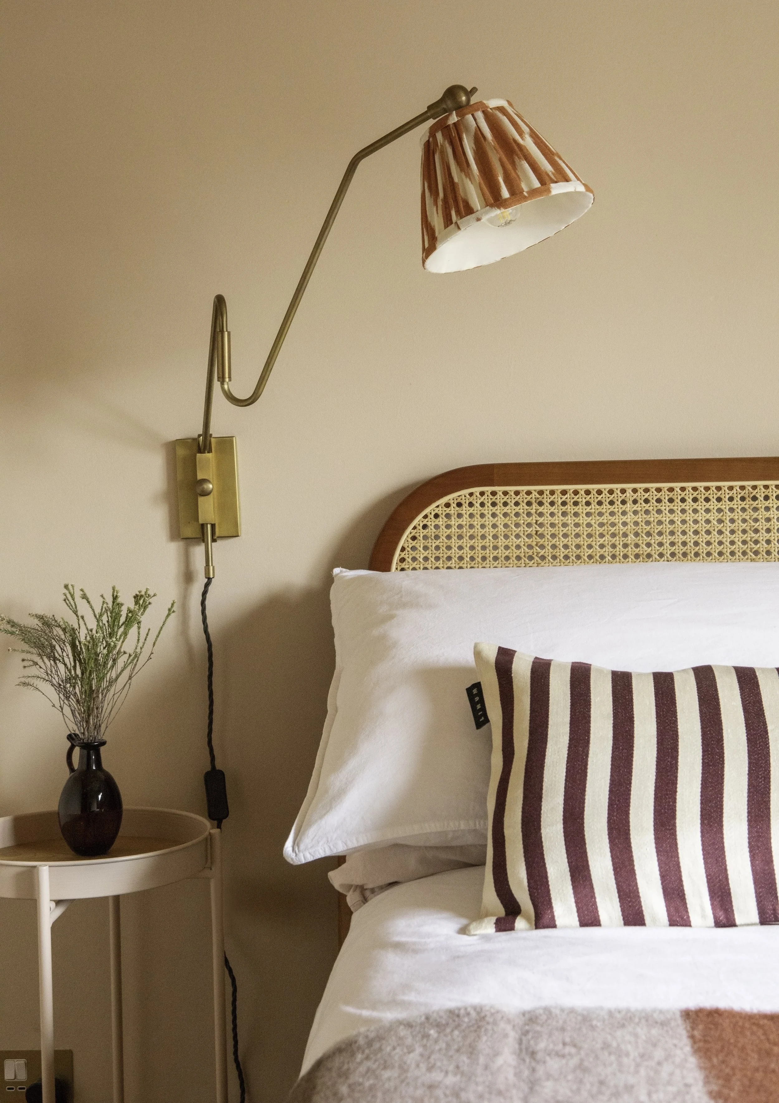 A bedroom with a bed, white pillow, and striped pillow. A wall-mounted lamp with a patterned lampshade and a small side table with a dark vase and green plant.