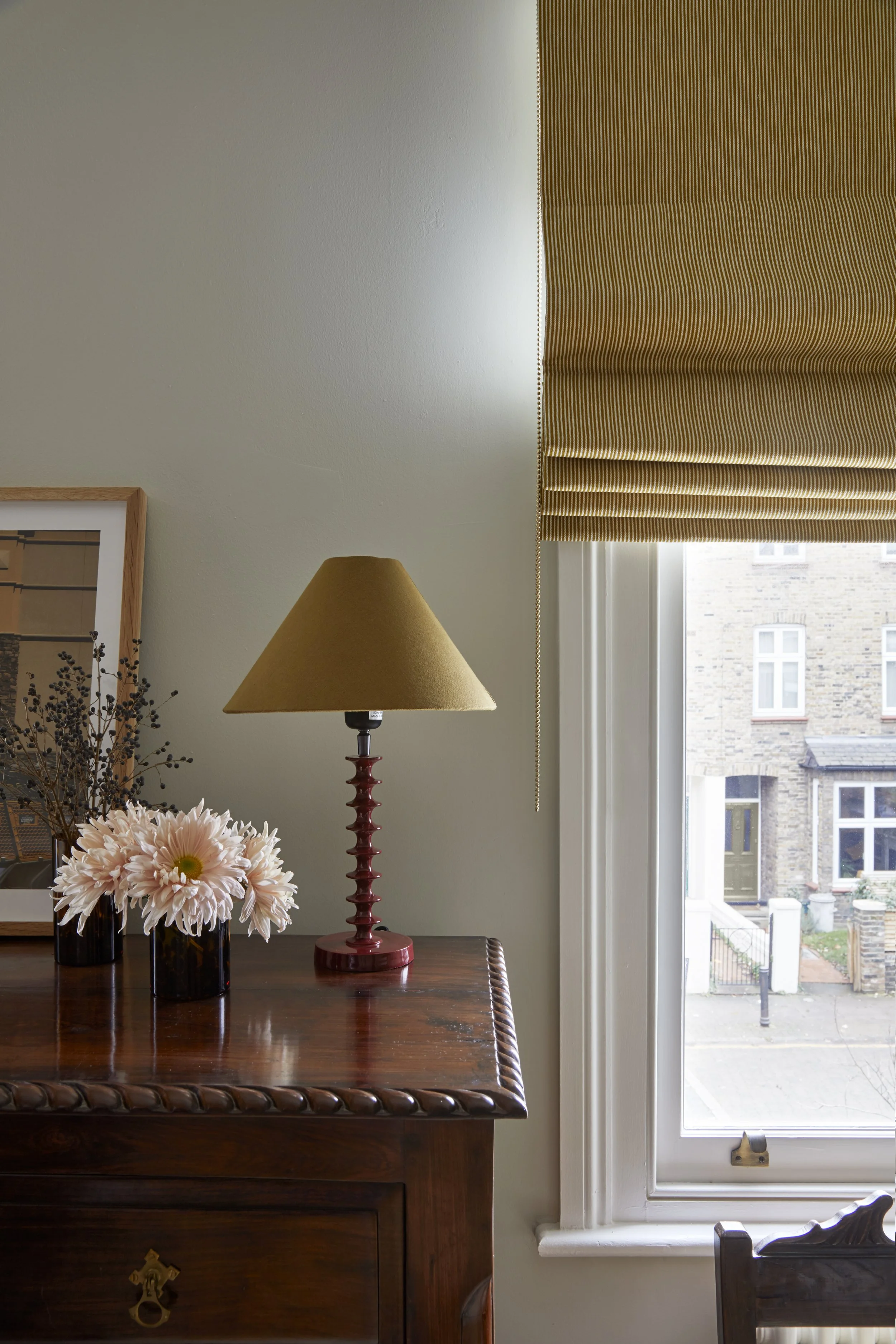 Large dresser , lampshade with a glossy metal red base, a vase of light pink flowers in a speckled vase, and a window with a golden-striped Roman blinds, 