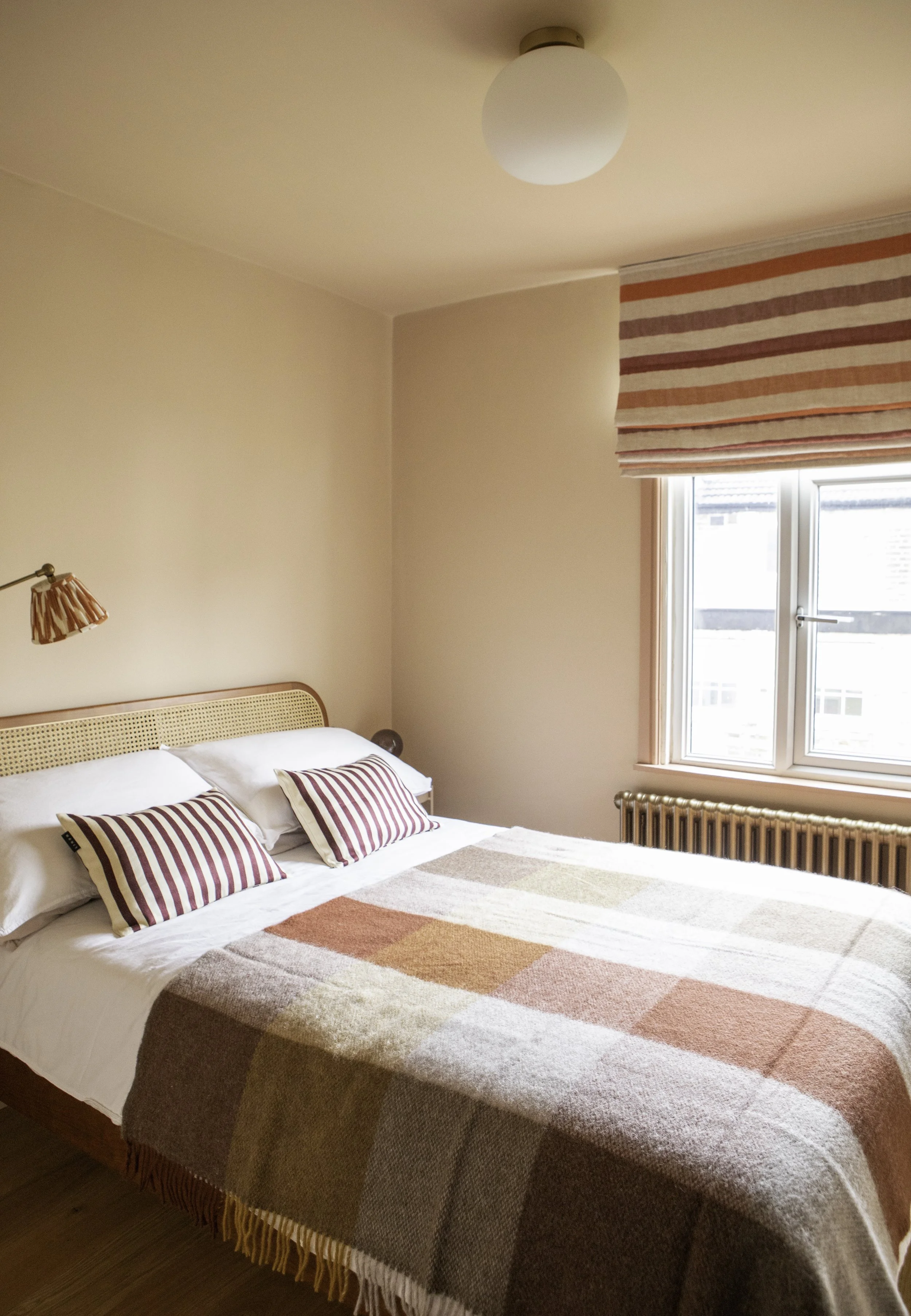 A cozy bedroom with a bed featuring striped pillows and a patchwork blanket, a window with a striped Roman shade, a wall-mounted reading lamp, and a ceiling light fixture.