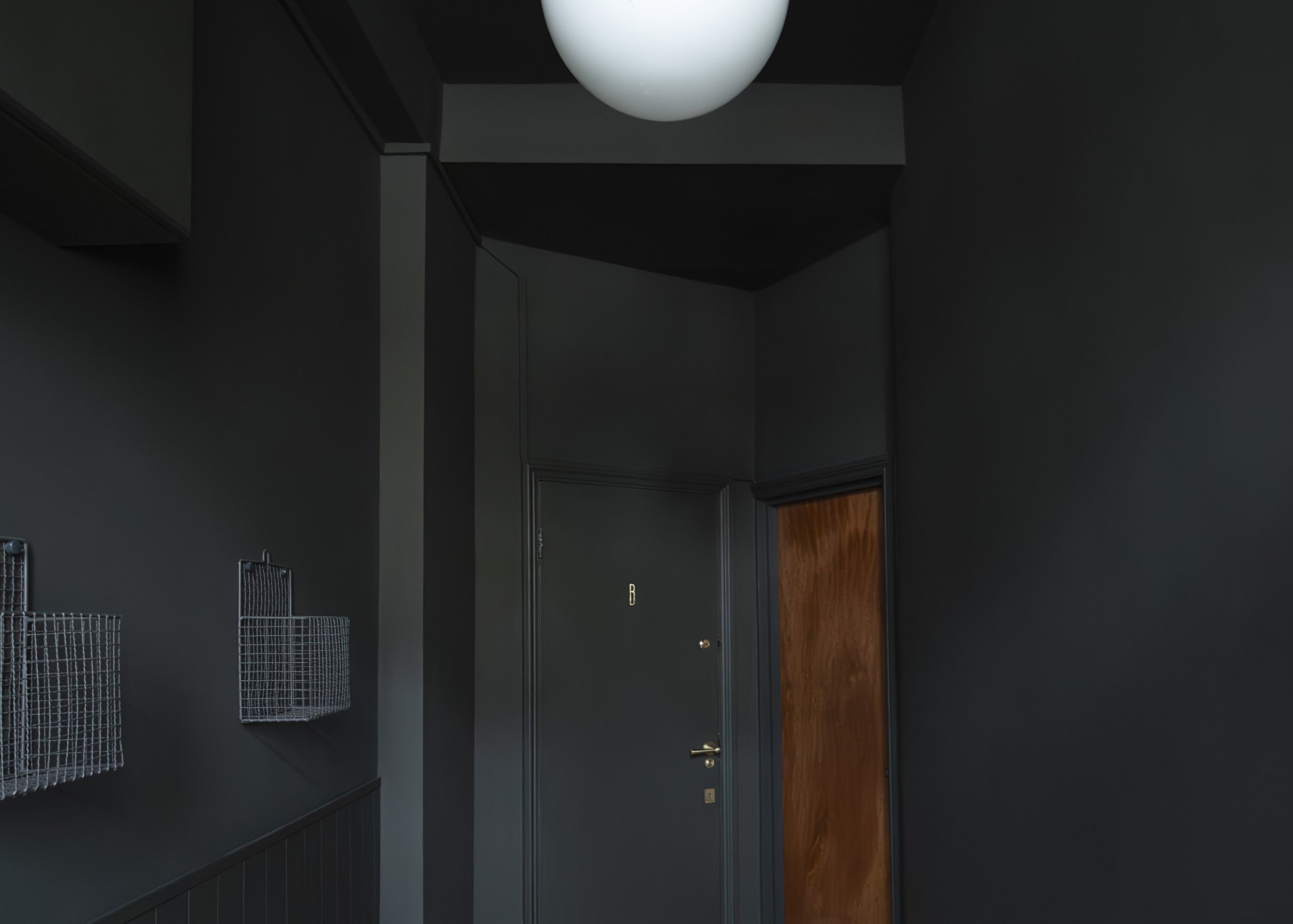 Dark hallway with green walls, a white vintage spherical ceiling light, metal wire baskets mounted on the wall, a green door marked with a letter B in brass, and a wooden door.