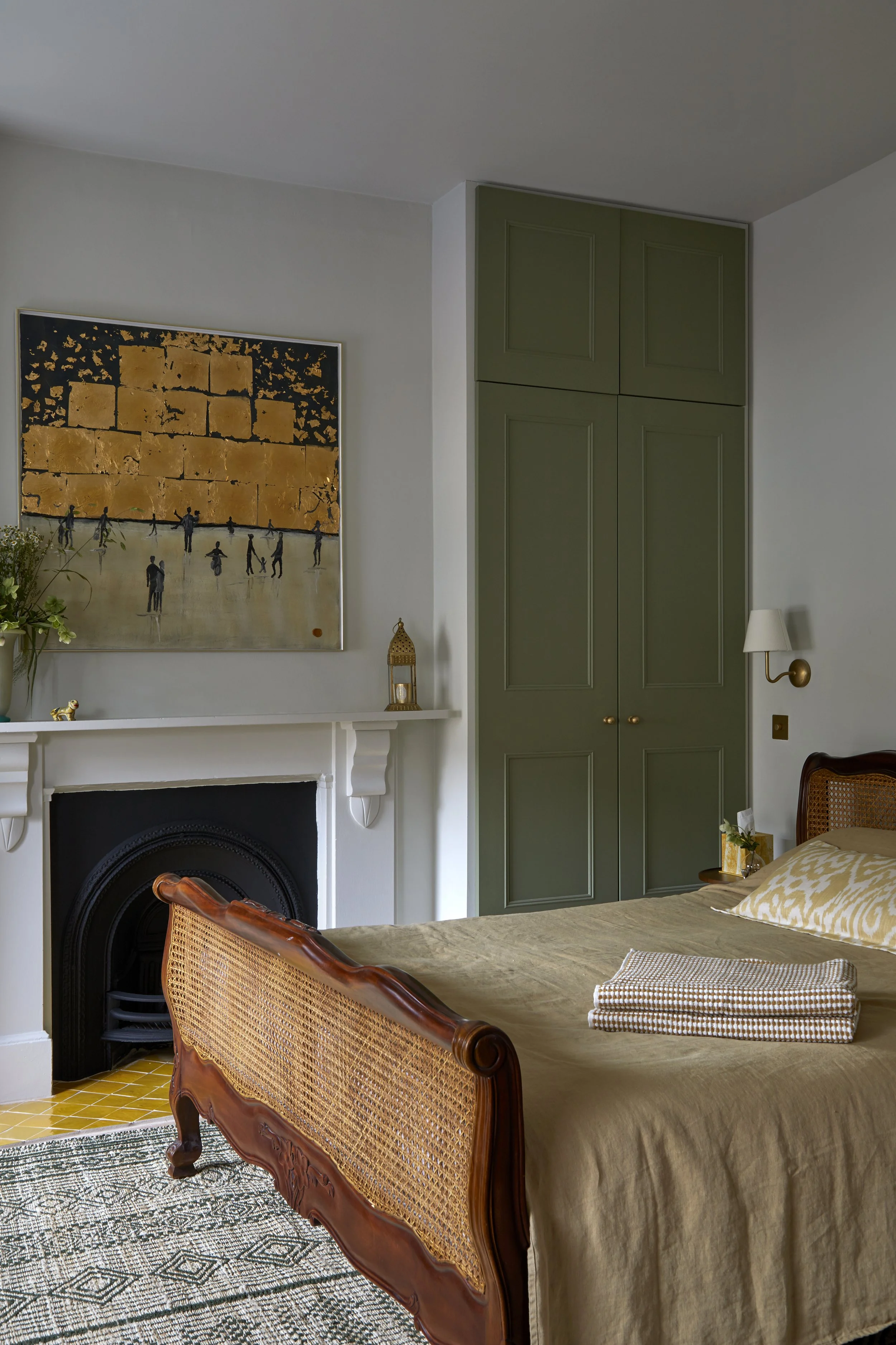 A bedroom with painted green wardbrobes , a painting above the fireplace, a wooden bed with a rattan headboard, and a folded towel on the bed.