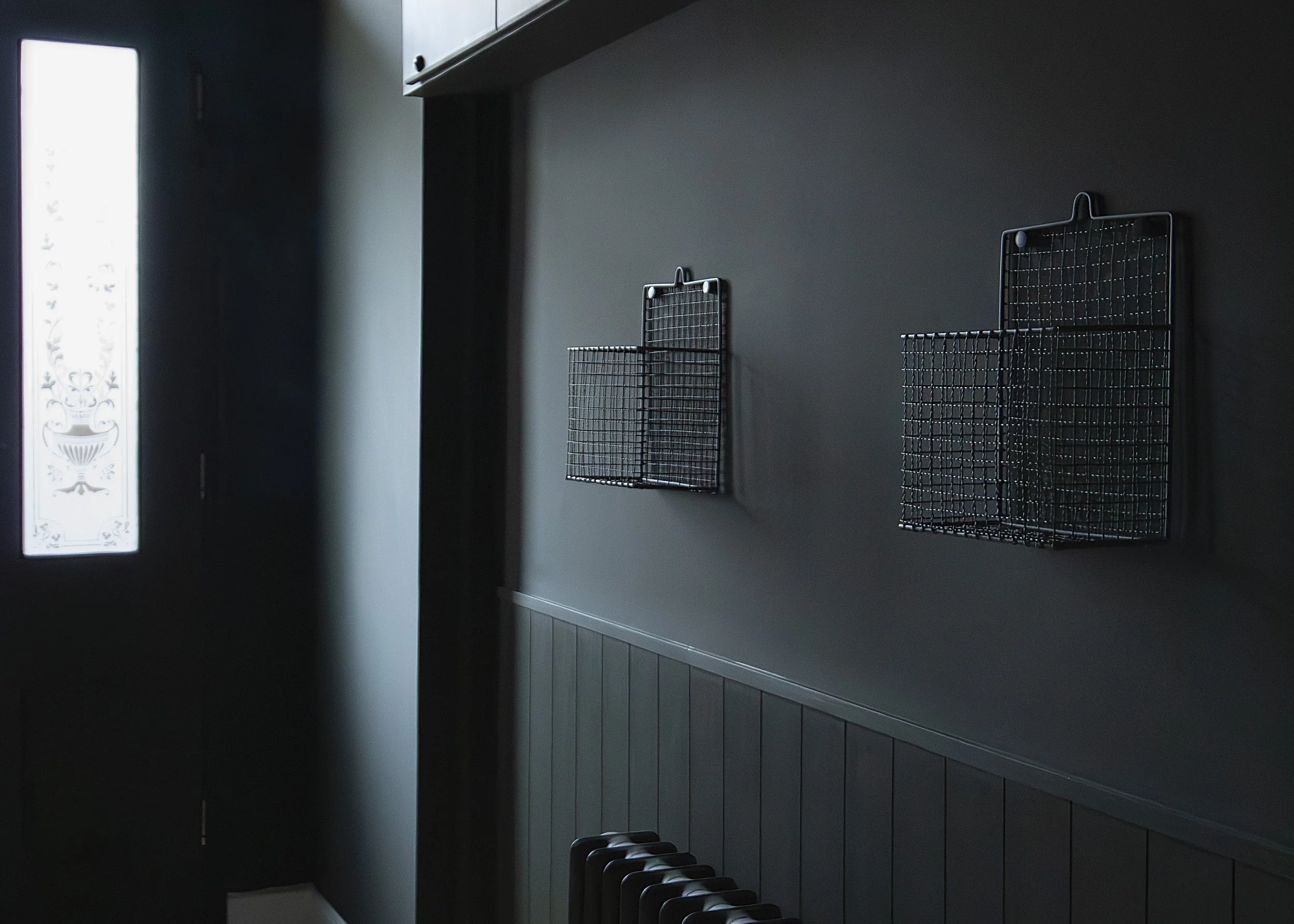 Dark green wall with two metal wire baskets hanging, one small and one larger, near a window with frosted glass and decorative pattern.