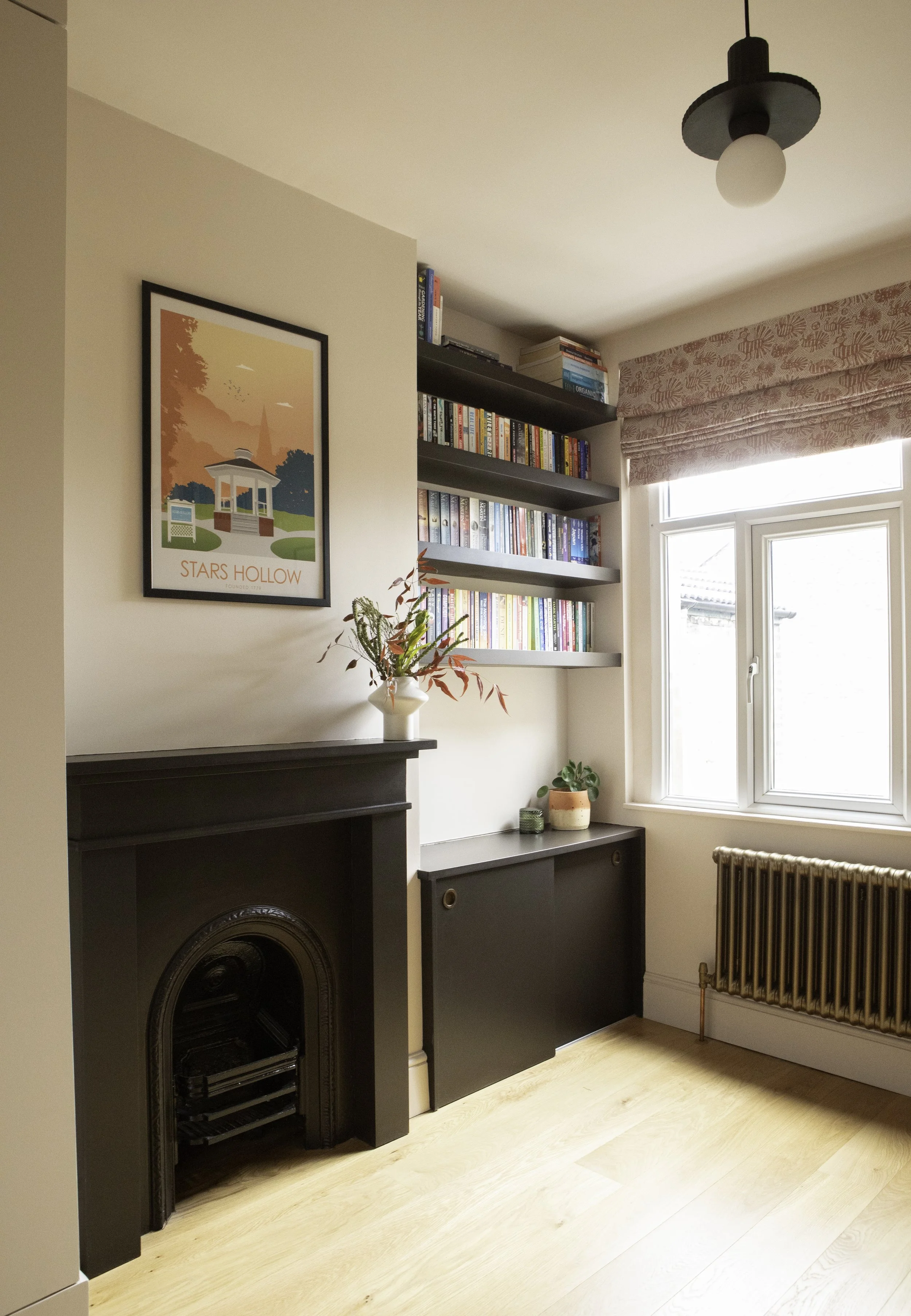 A cozy living room corner featuring a black fireplace mantle, a white vase with red and green foliage on top, a black bookshelf filled with colorful books, a window with pink patterned Roman blinds, and a bronze radiator below the window.