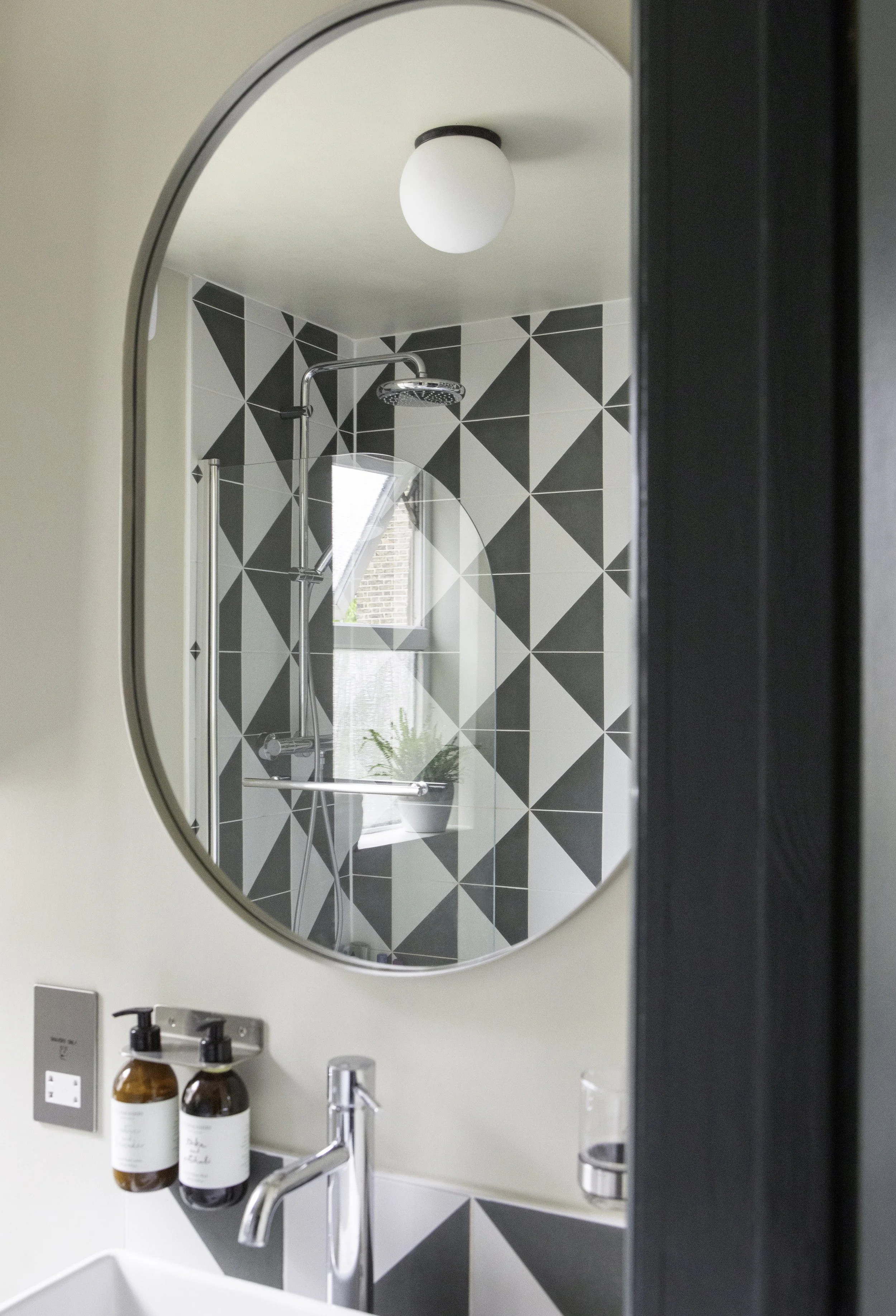 Bathroom mirror reflects a shower area with geometric patterned tiles and a small window with a potted plant. The sink area has two soap dispensers and a modern faucet.