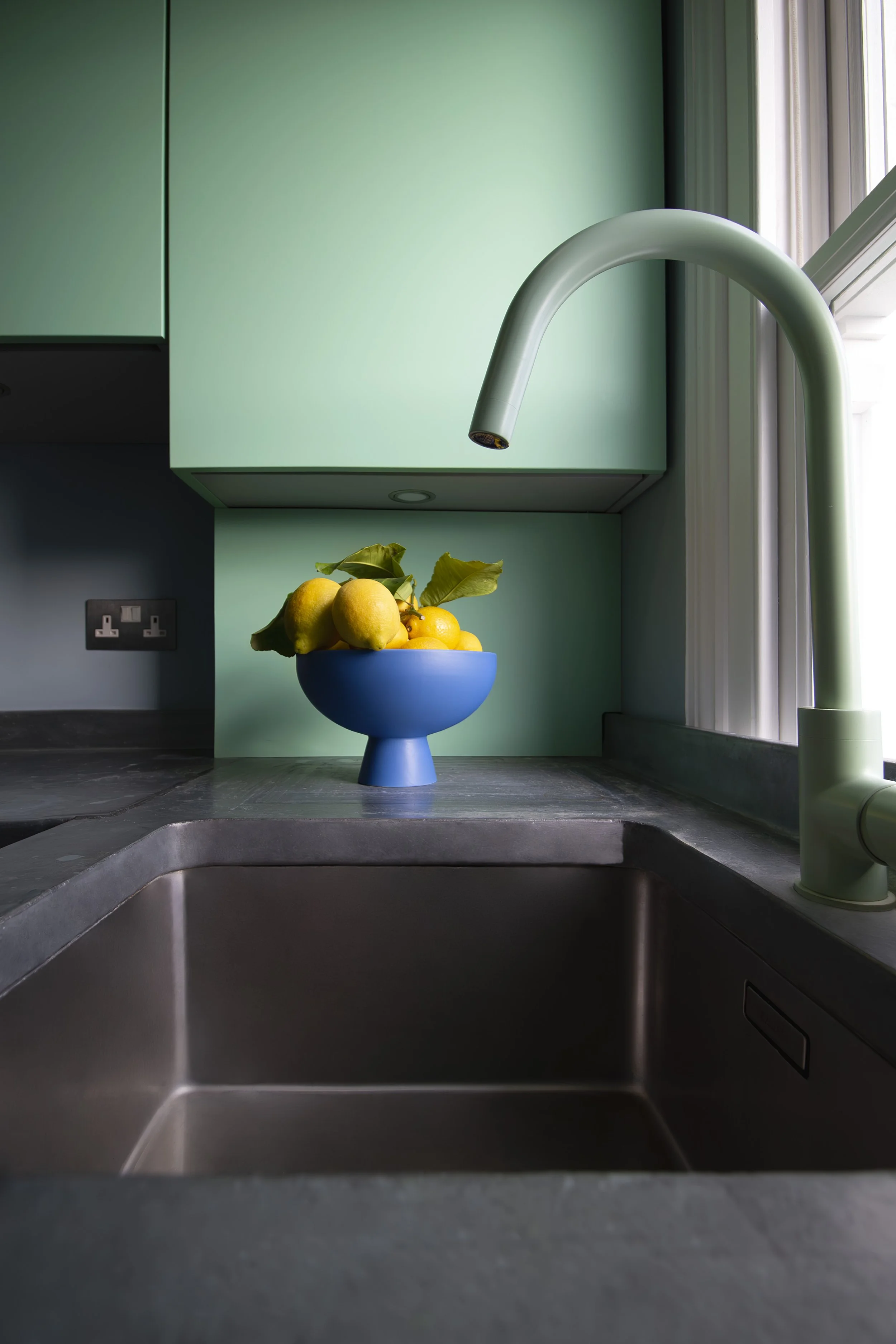 A kitchen sink area with a modern green cabinet, a white faucet, a bowl of lemons, and a window with curtains.