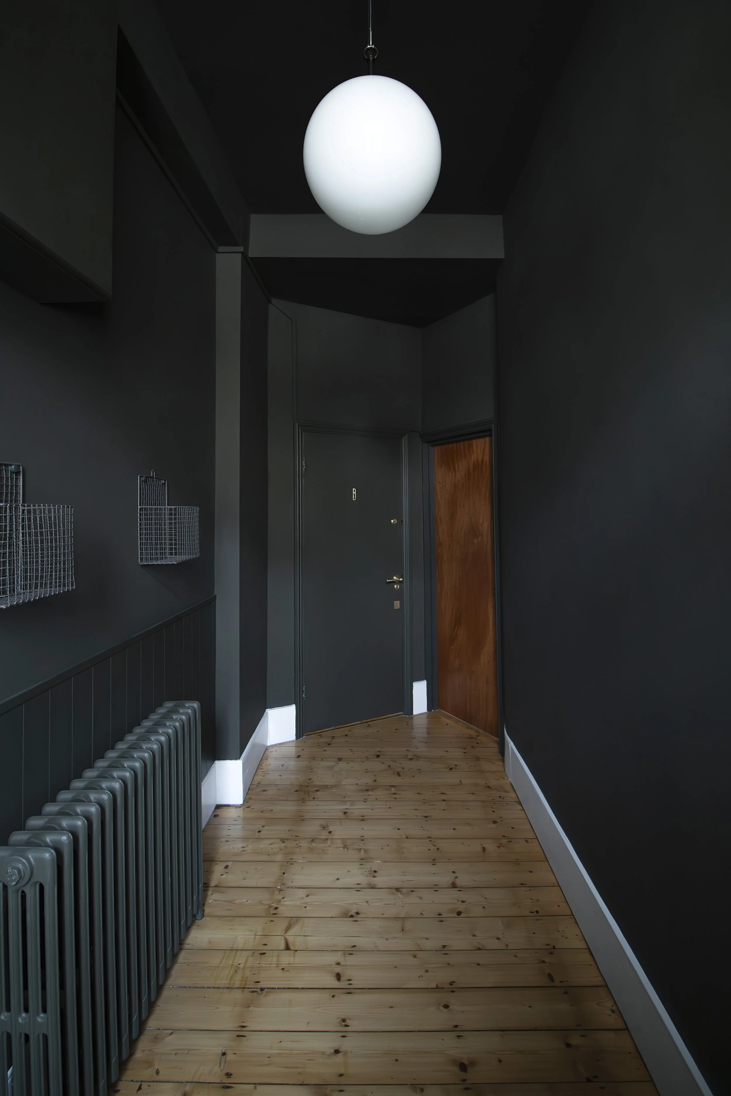 Dark hallway with green walls, a wooden floor, a black front door with the letter 'B', a brown door, a modern white pendant light, a green radiator, and black wire wall baskets.