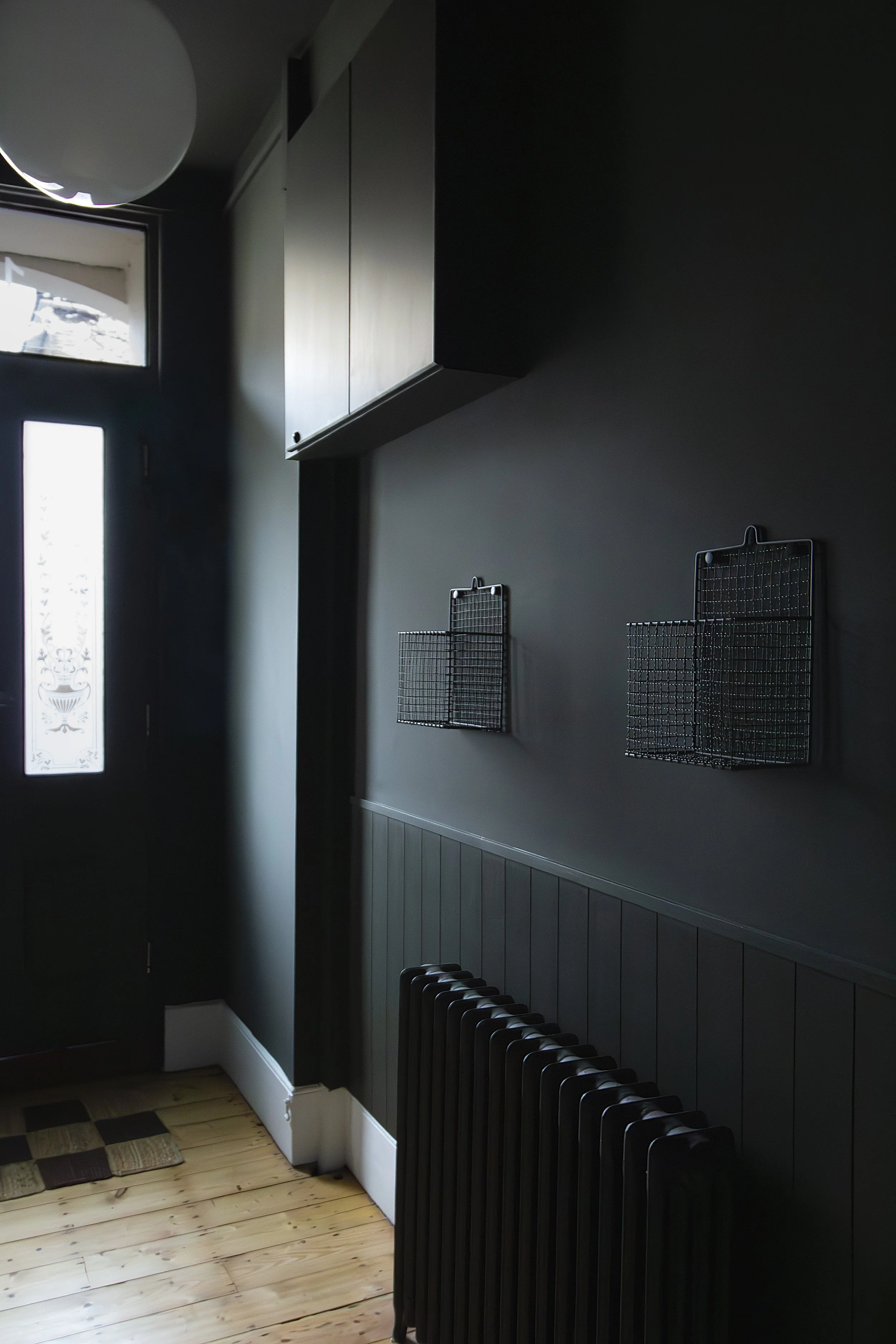 Dark-colored indoor corner with a door, wall-mounted wire baskets, a radiator, and a wood floor.