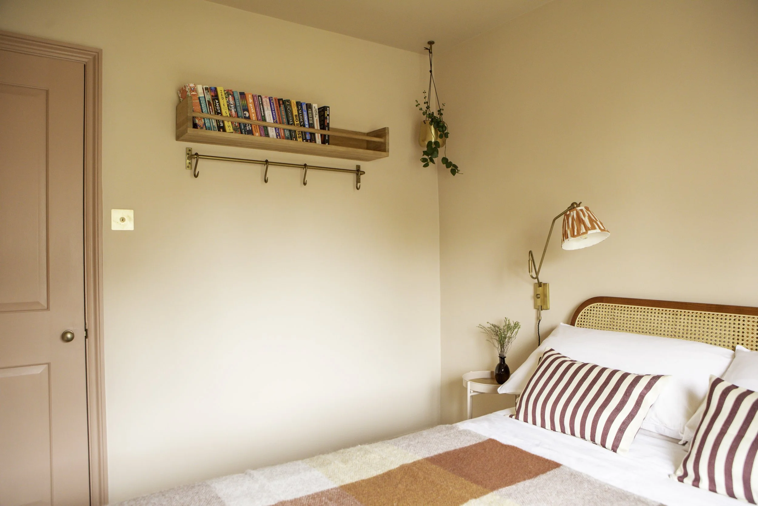 A cozy bedroom with a bed, striped pillows, a bedside table with a small plant, a wall-mounted lamp, a floating bookshelf filled with books, a hanging plant, and a peg hook rod on the wall.