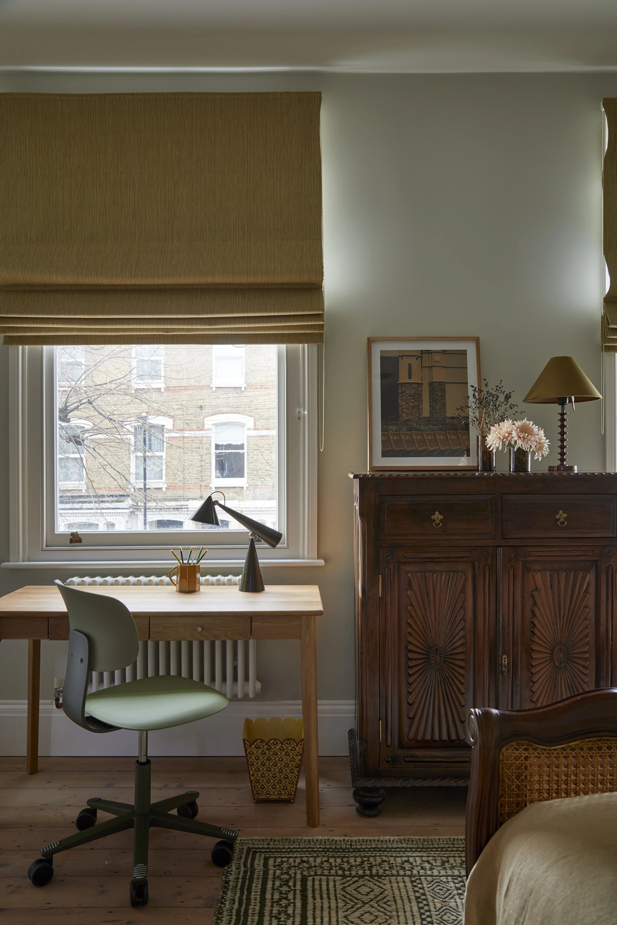 A cozy home office with a wooden desk, green swivel chair, tom dixon desk lamp, window, wooden cabinet with framed artwork and flowers, roman blinds in Liberty Fabric, and a patterned rug.