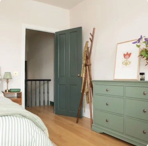 A bedroom with a green door partially open, a green dresser, a framed floral art on the wall, a vase with flowers, and a wooden coat rack leaning against the wall.