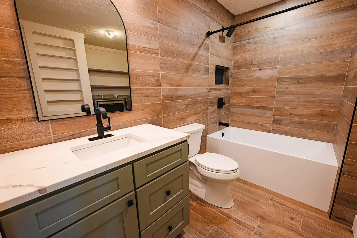 Spa style bathroom with green vanity with white quartz counter, wood style tile on floor and walls, white toilet and bathtub