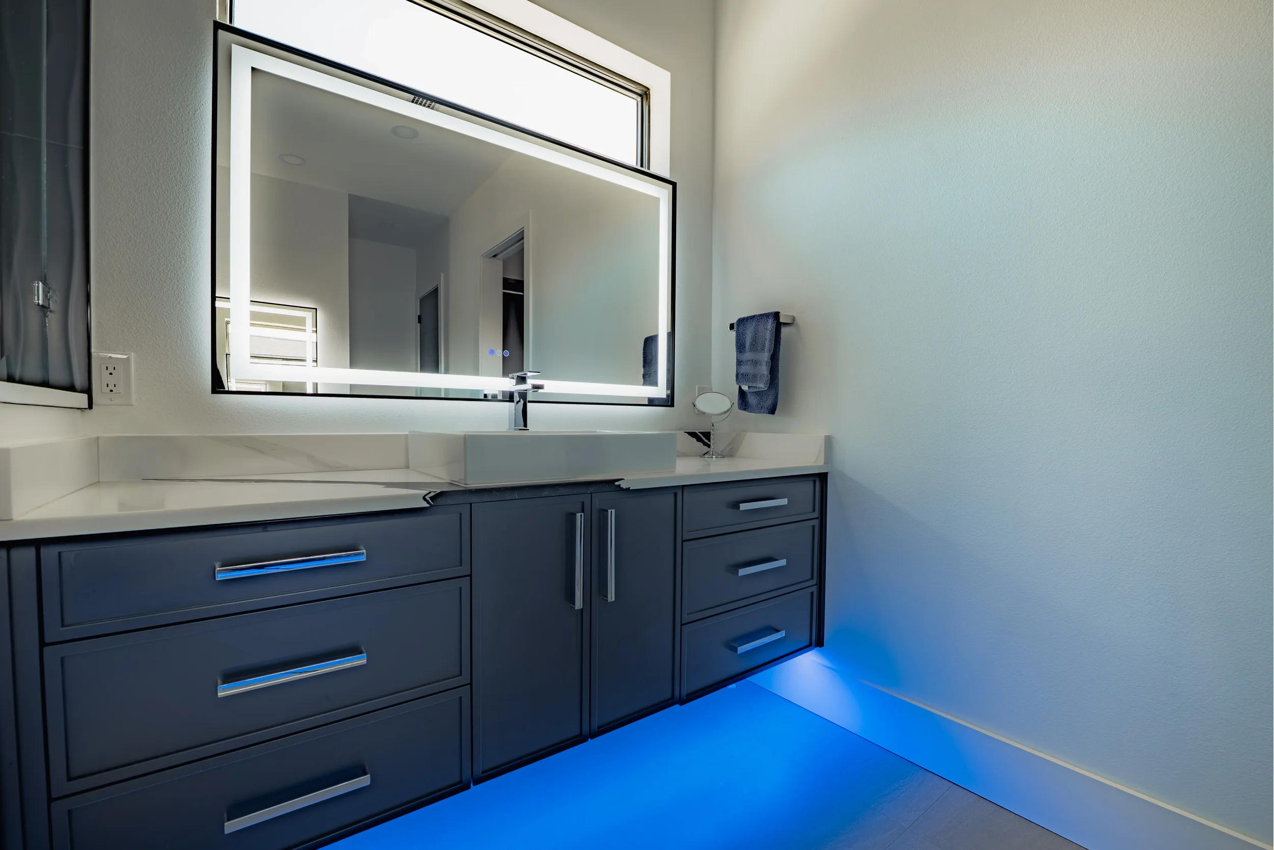 Black cabinet vanity floating with blue under glow lighting. Big mirror and ceramic sink with transom window above mirror.