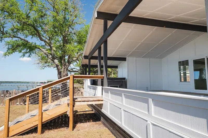 Rear view of a modern house with a wooden ramp, white siding, and an overhanging roof near a large green tree and a body of water in the background.