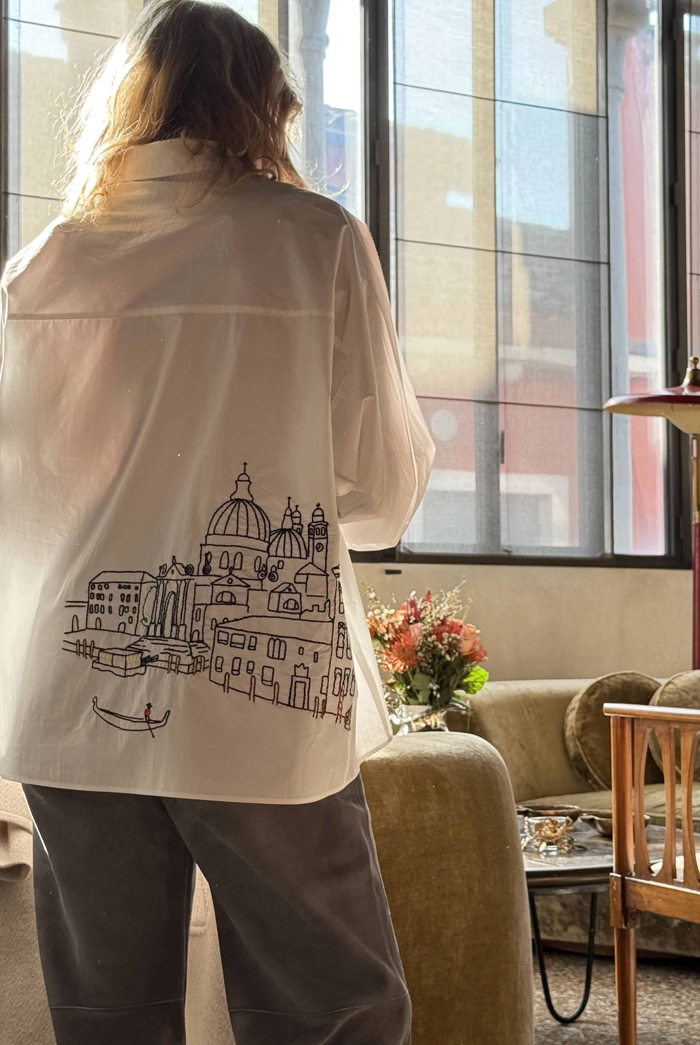 Person with long, curly hair wearing a white shirt with a cityscape and boat illustration, standing indoors near a large window with sunlight streaming in, with a flower arrangement and a sofa in the background.