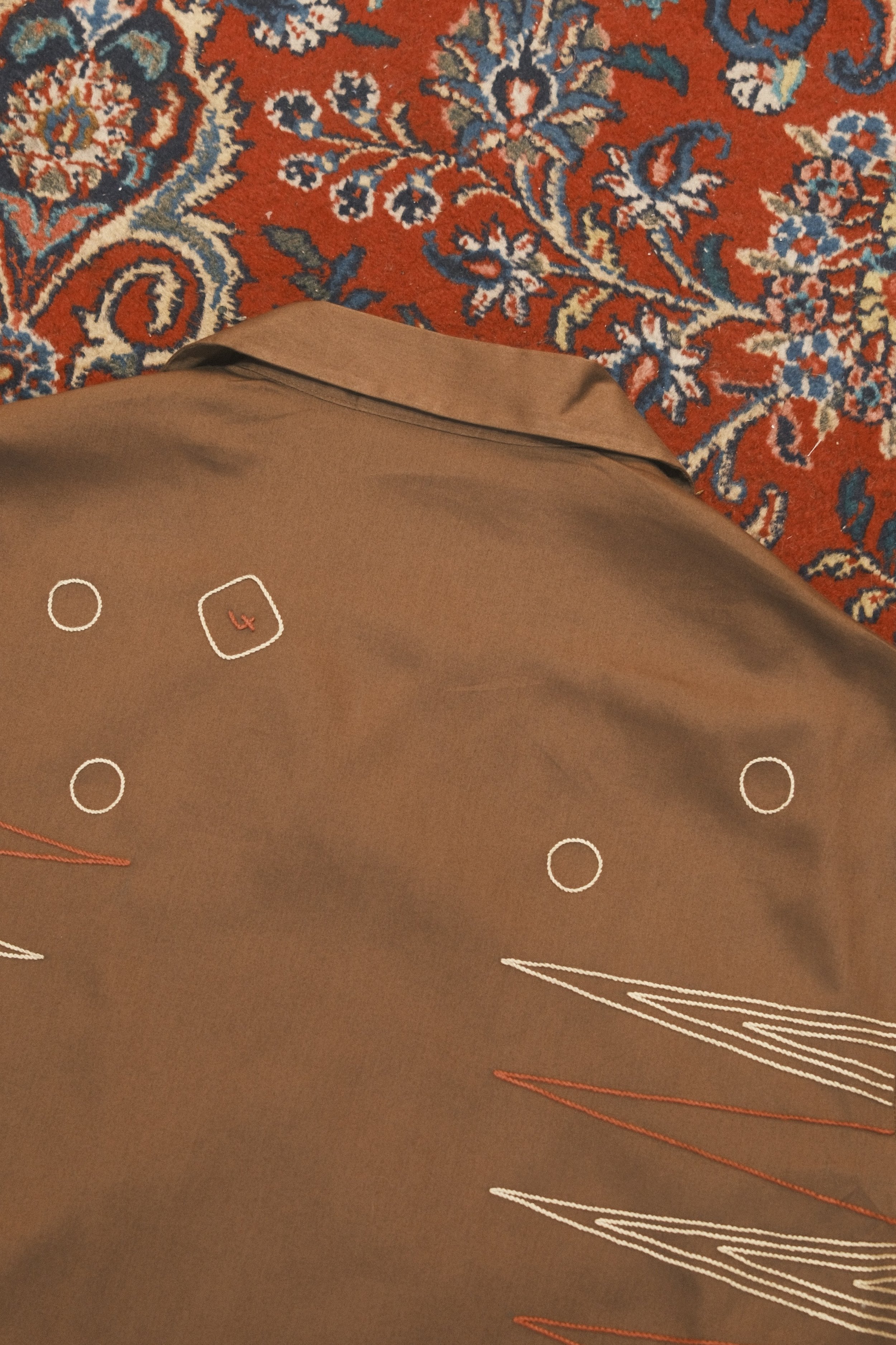 Part of a patterned rug on the floor and a person's brown pants with button details and embroidery, standing partially over the rug.