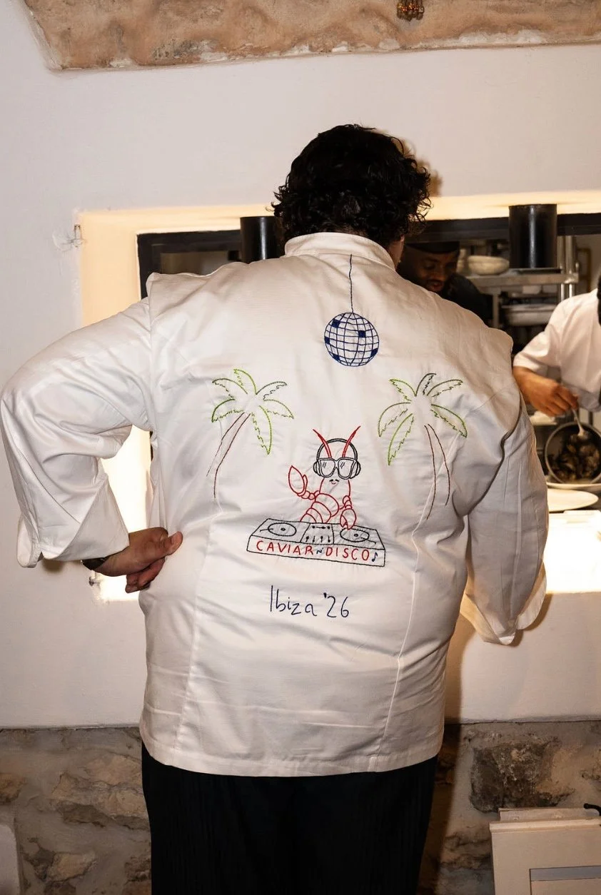Person wearing a white jacket with a colorful embroidered design of a lobster DJ wearing sunglasses, palm trees, a disco ball, and the text 'CAVIAR AND DISCO Ibiza '26' at the back.