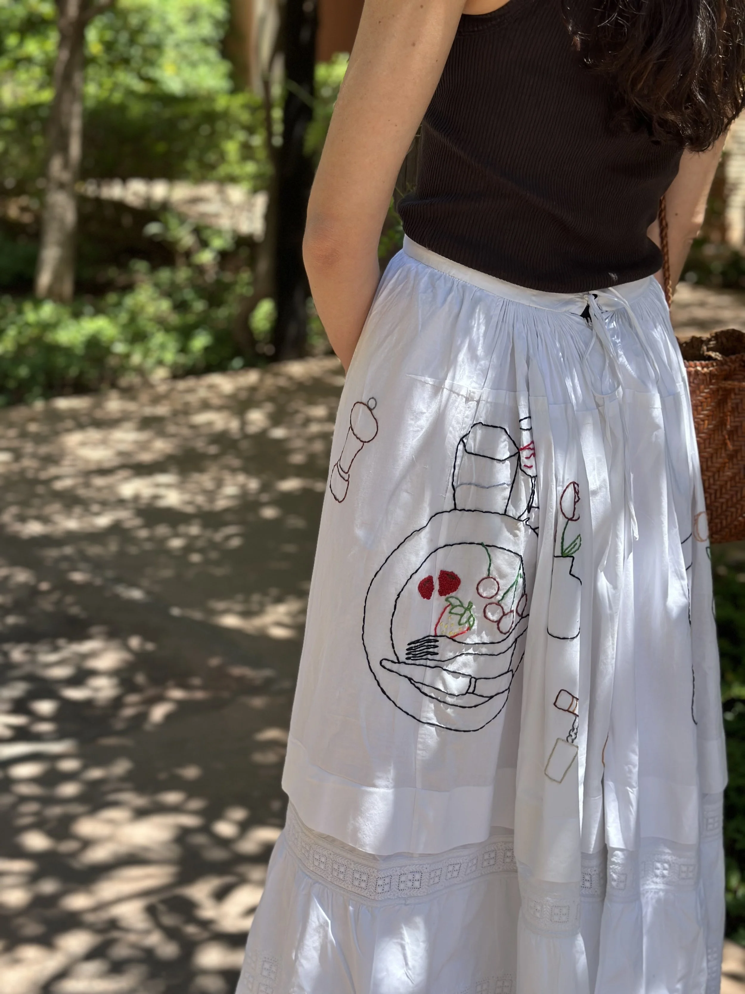 Close-up of a woman wearing a black sleeveless top and a white skirt with embroidered designs, standing outdoors with sunlight filtering through trees.