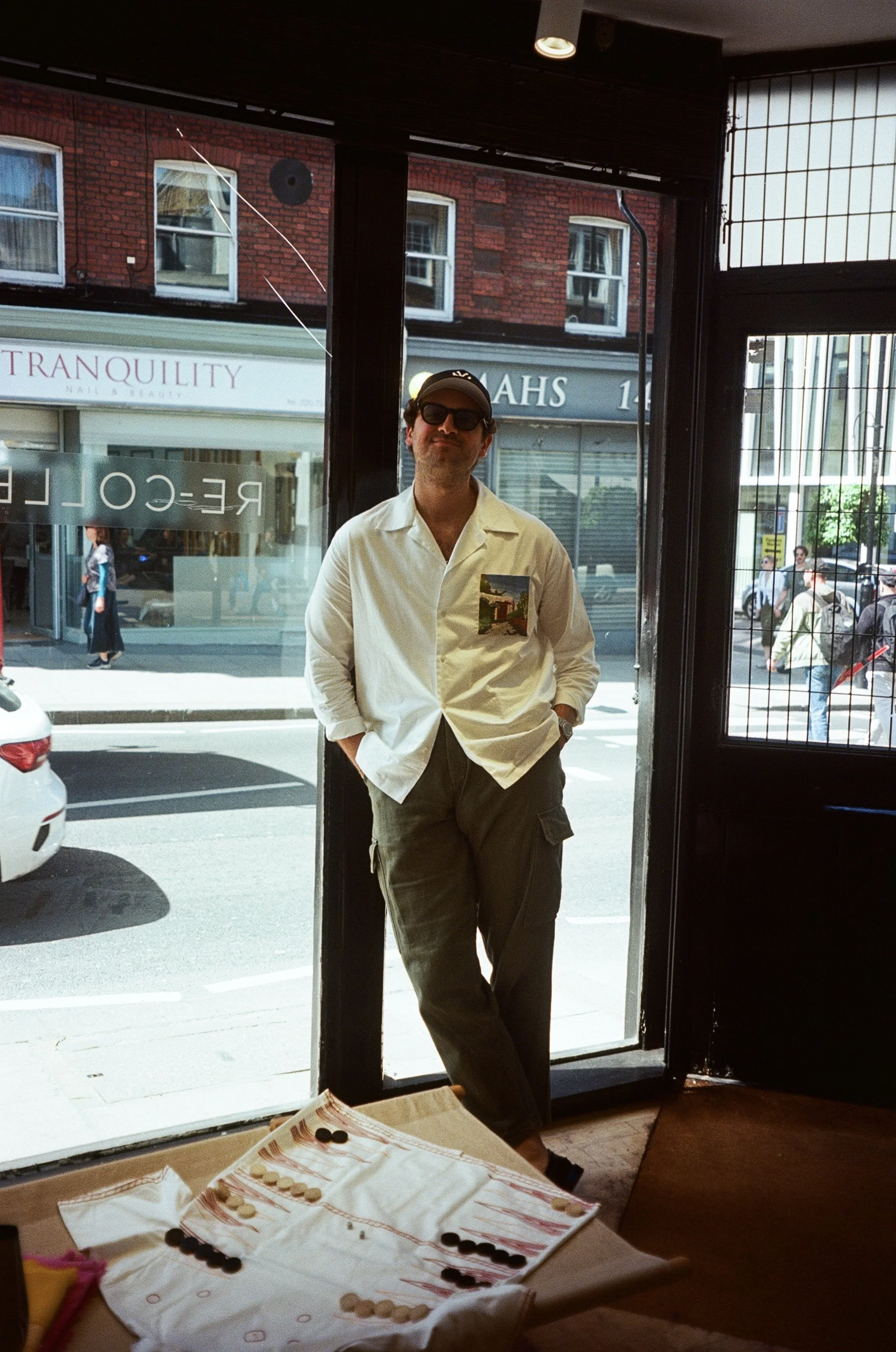A man wearing sunglasses, a white shirt with a sculpture print pocket, and cargo shorts, standing casually with hands in pockets outside a storefront on a sunny day.