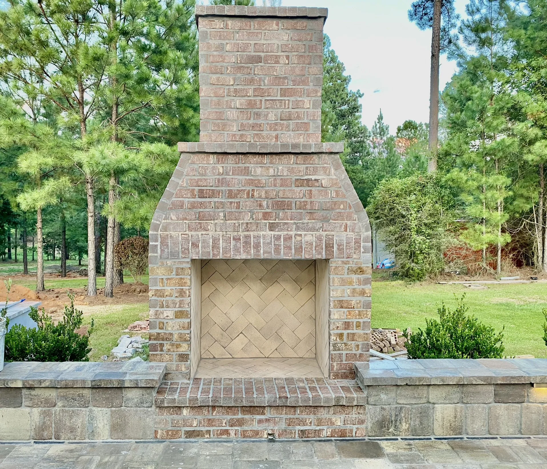 Outdoor brick fireplace with a herringbone interior design, set in a landscaped area with lush green trees in the background.