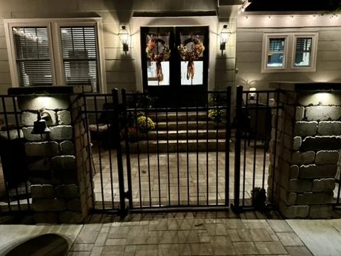Front porch with a gated entry, illuminated at night, featuring stone pillars, fall wreaths on the door, and warm outdoor lighting.