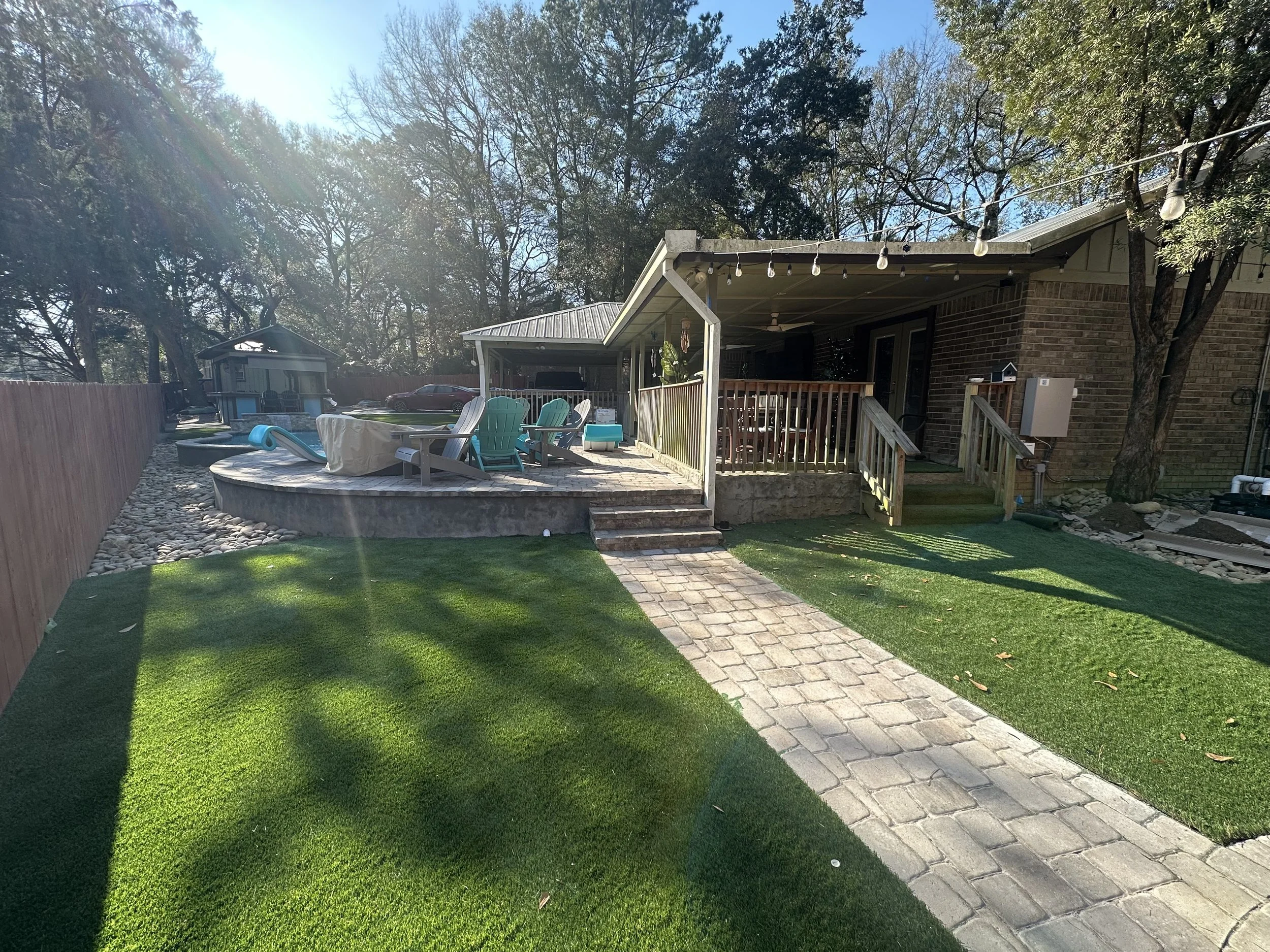 Inviting Gulf Coast backyard with artificial turf, paver walkway, elevated patio seating area, and string lights for outdoor relaxation.