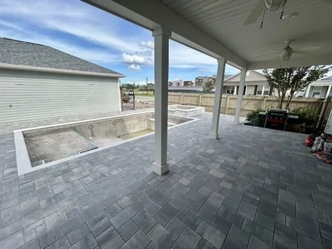 Covered patio with gray stone flooring and ceiling fans, overlooking an unfinished in-ground pool and a fenced backyard on a clear day.