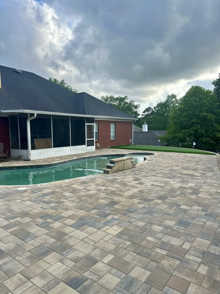 Backyard featuring a stone-paved patio surrounding an in-ground pool, with a red brick house and screened porch under a cloudy sky.