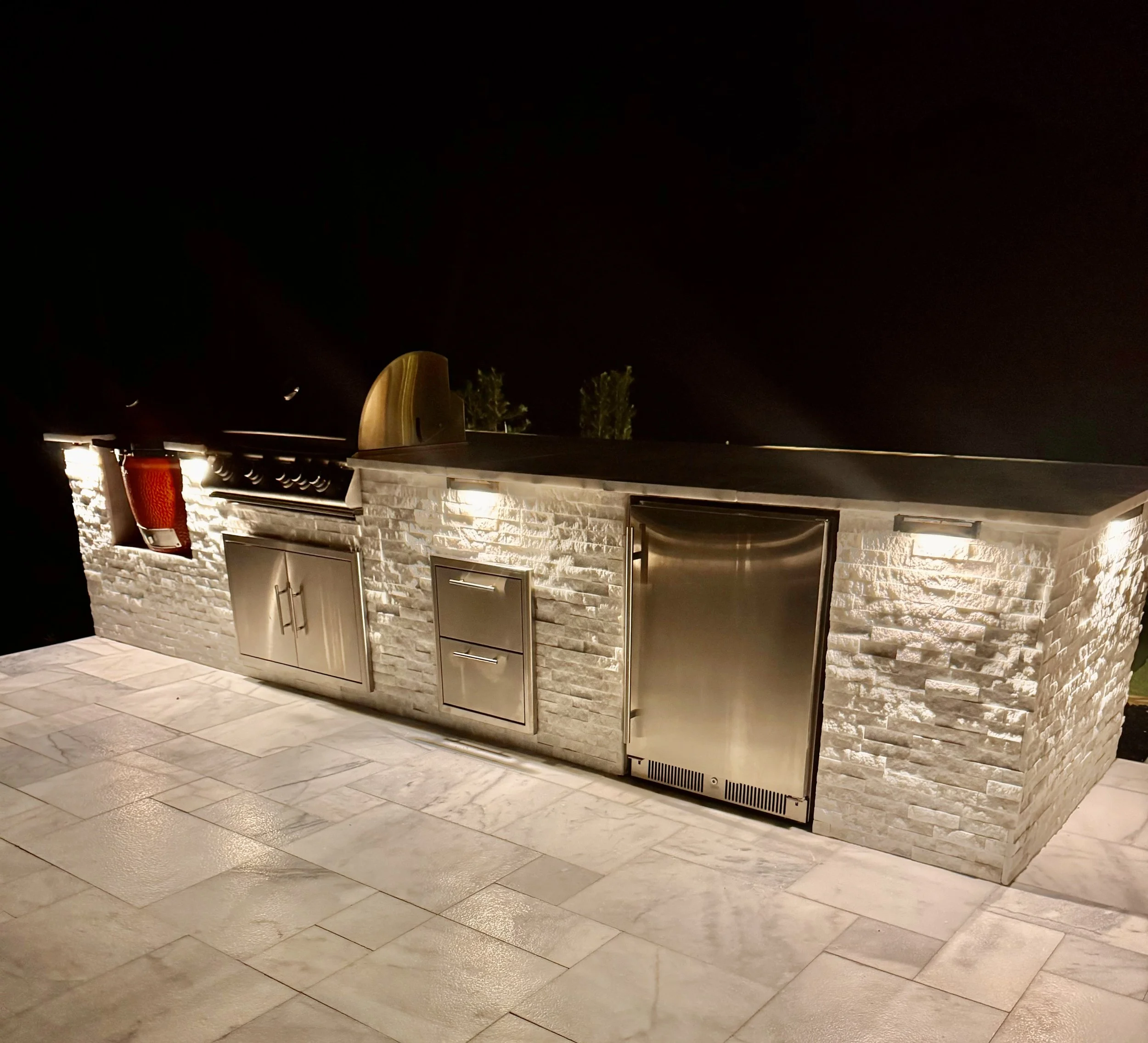 Night view of an outdoor kitchen with a stone facade, stainless steel appliances, and a lighted countertop on a tiled patio.