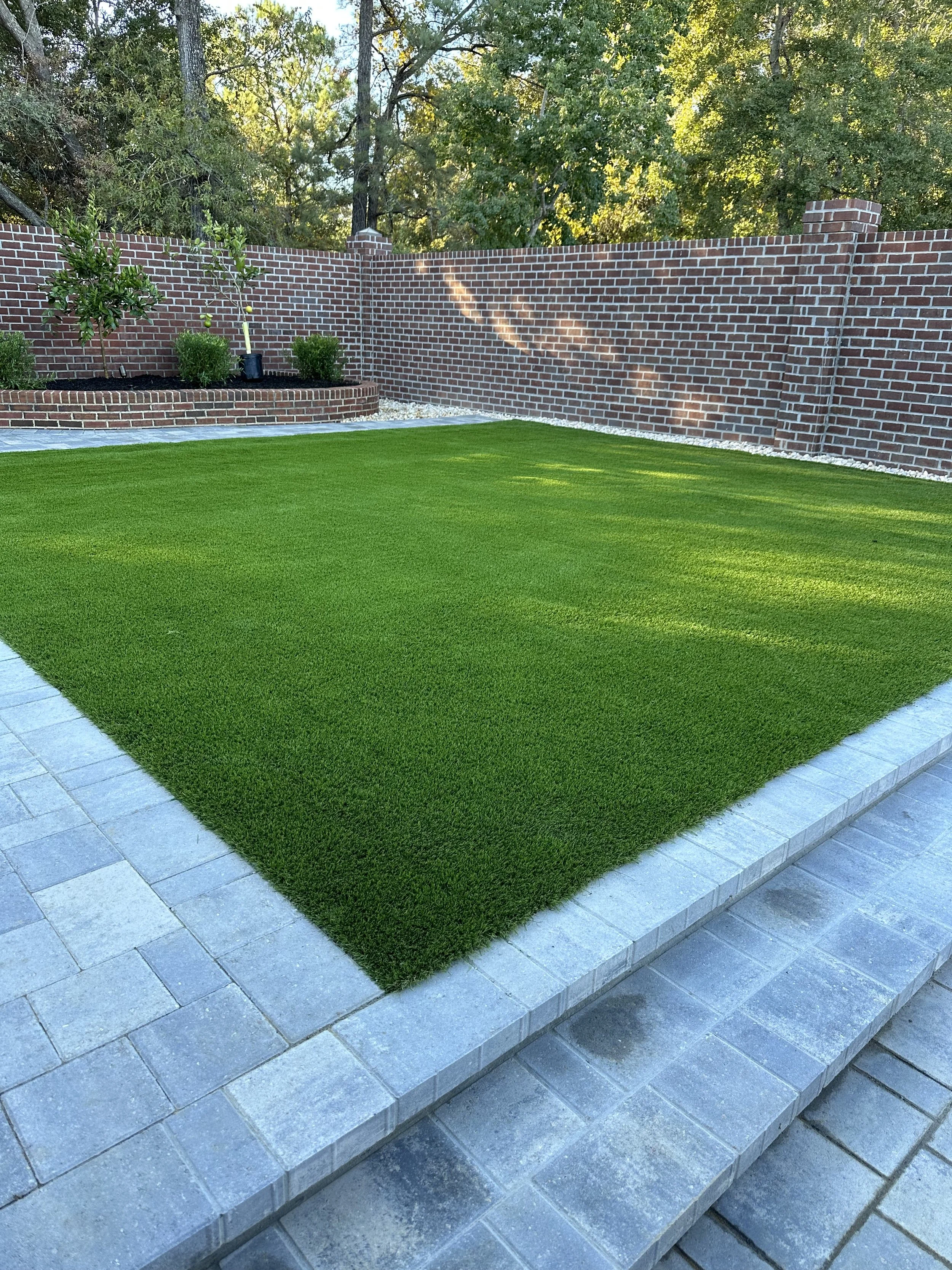 Backyard with green artificial grass, gray stone pavers, brick wall, and small trees.