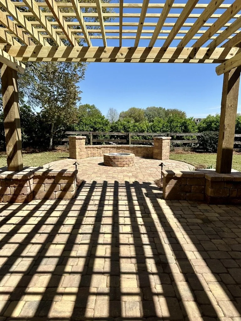 A scenic outdoor patio with a wooden pergola casting shadows on a paved stone floor, featuring a circular fire pit and stone seating surrounded by lush greenery under a clear blue sky.