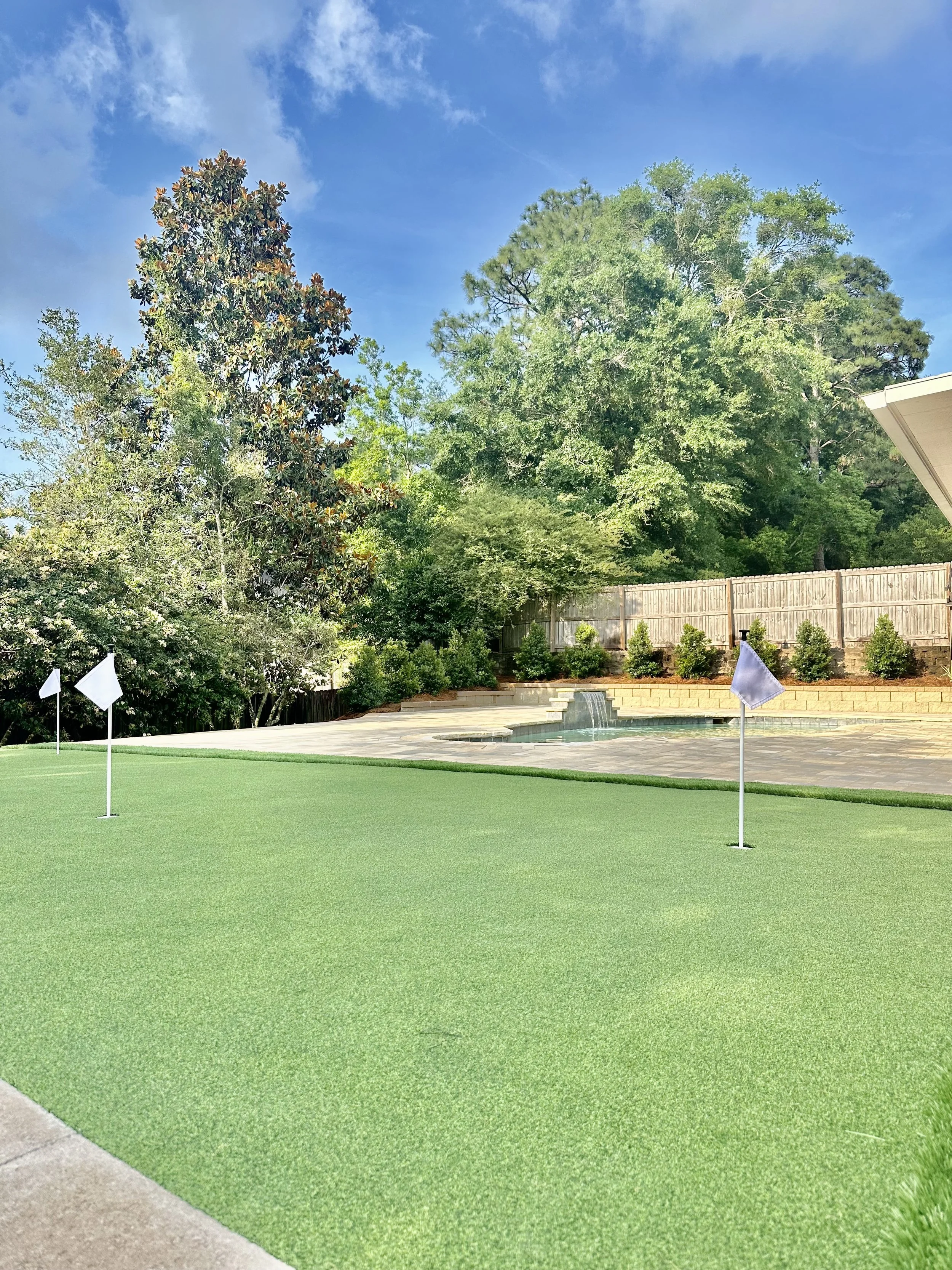 Backyard putting green with artificial turf next to a pool in a Gulf Coast landscape, designed for relaxation and golf practice.