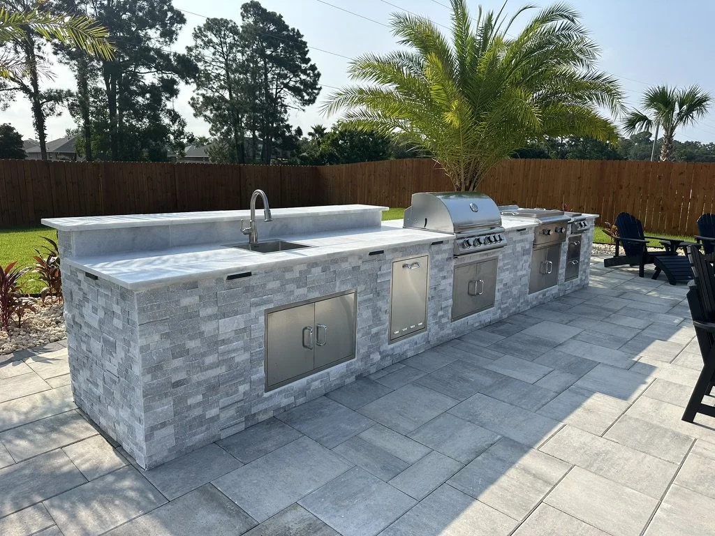 Modern outdoor kitchen with a stone countertop, built-in stainless steel grill, sink, and storage cabinets, set on a paved patio with palm trees and a wooden fence in the background.