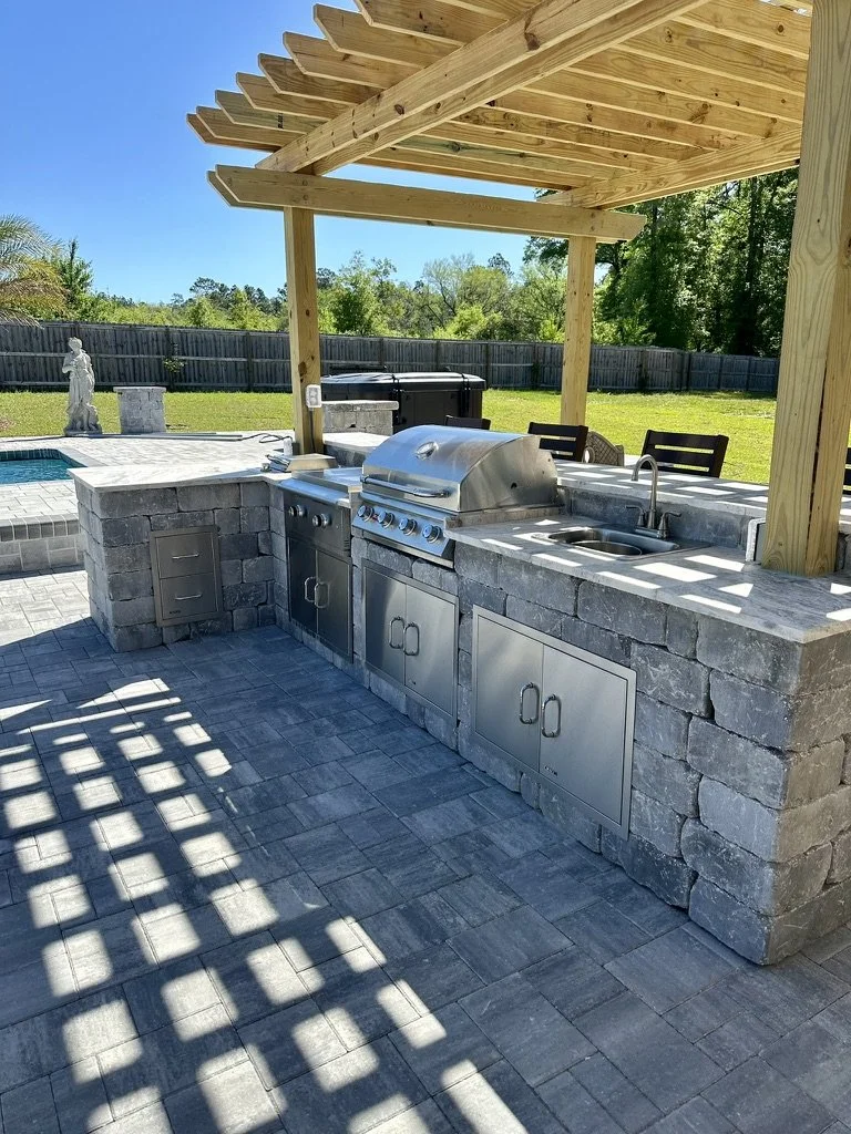Modern outdoor kitchen with pergola, stainless steel appliances, and stone pavers.