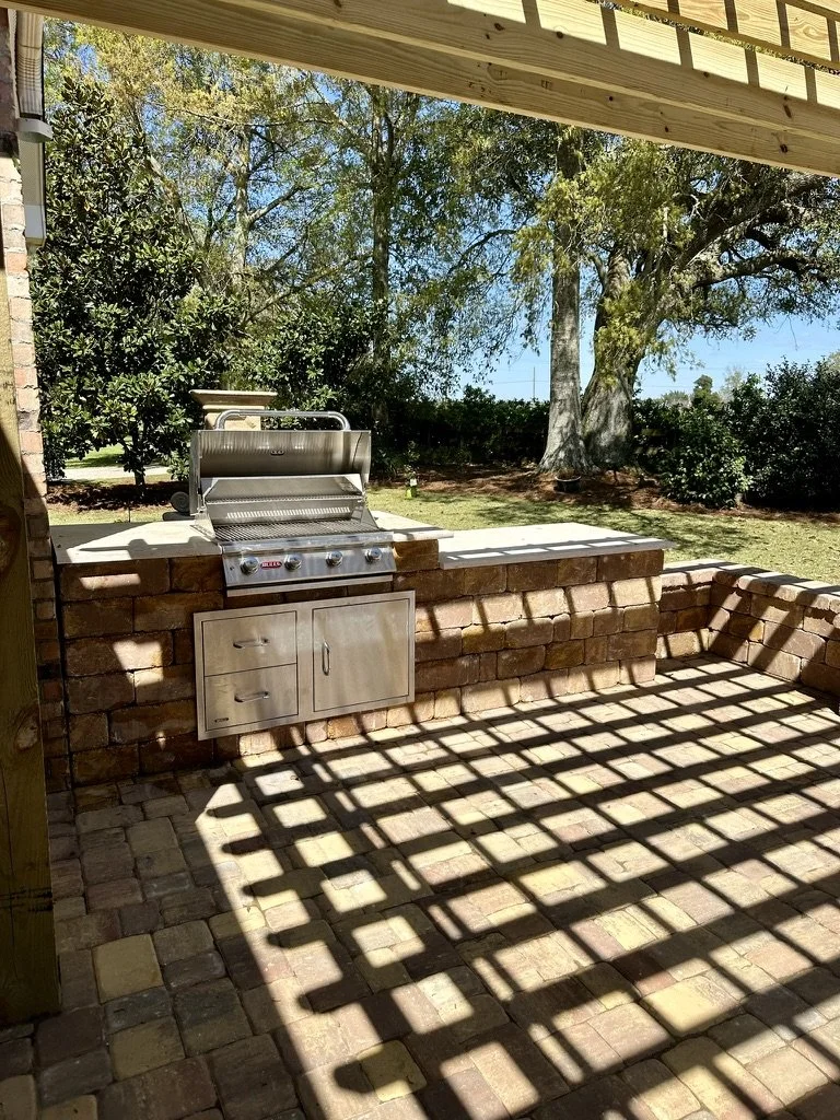 Outdoor kitchen area with a built-in stainless steel grill and stone wall, shaded by a wooden pergola casting patterned shadows on the paved patio.