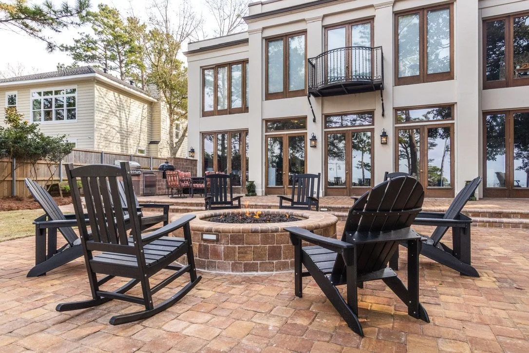 Luxury patio with a central fire pit surrounded by black Adirondack chairs, set against a modern multi-story home with large windows and a balcony.
