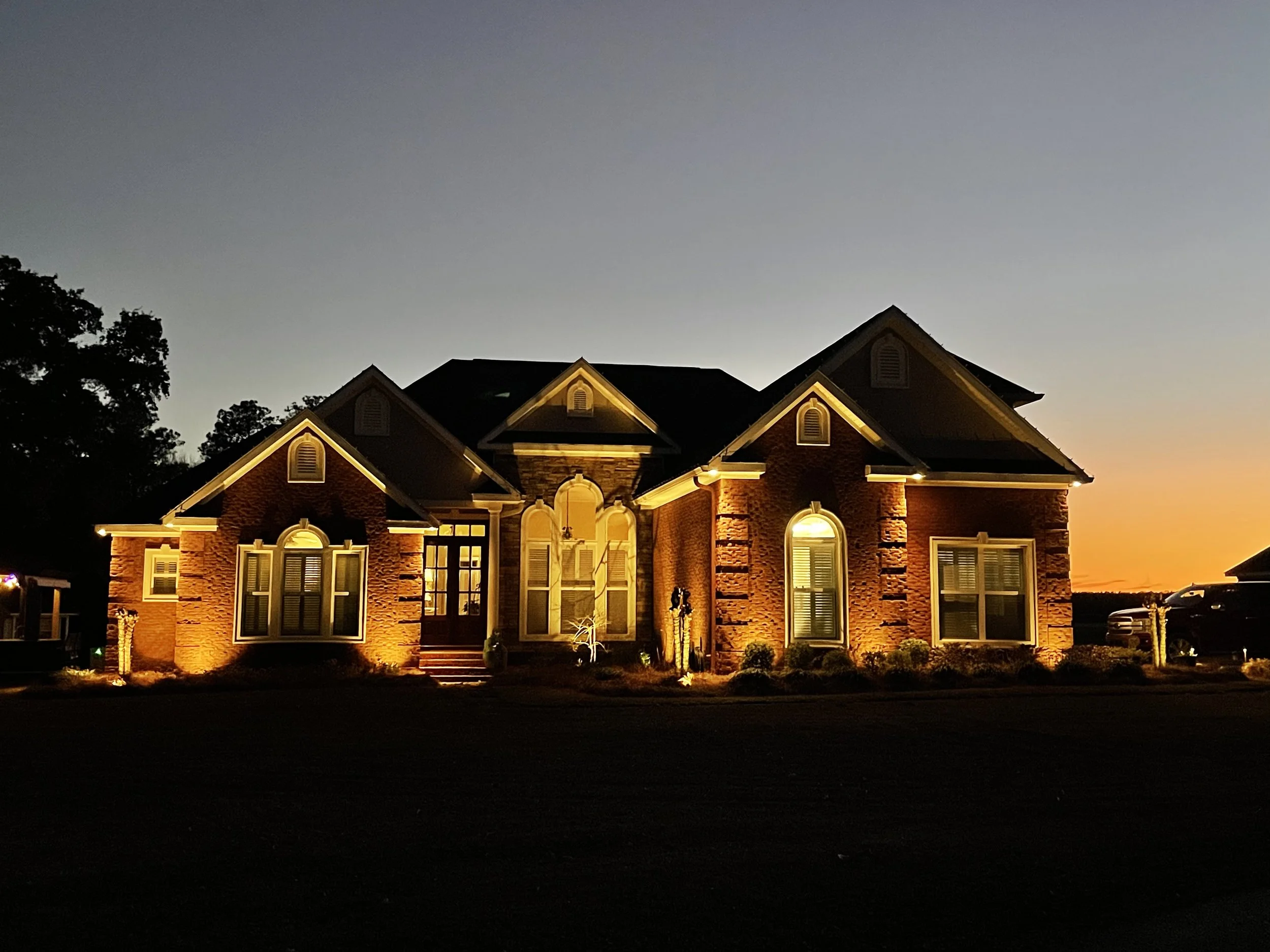 Brick home at sunset with landscape lighting accentuating windows, entryway, and surrounding gardens in a Gulf Coast setting