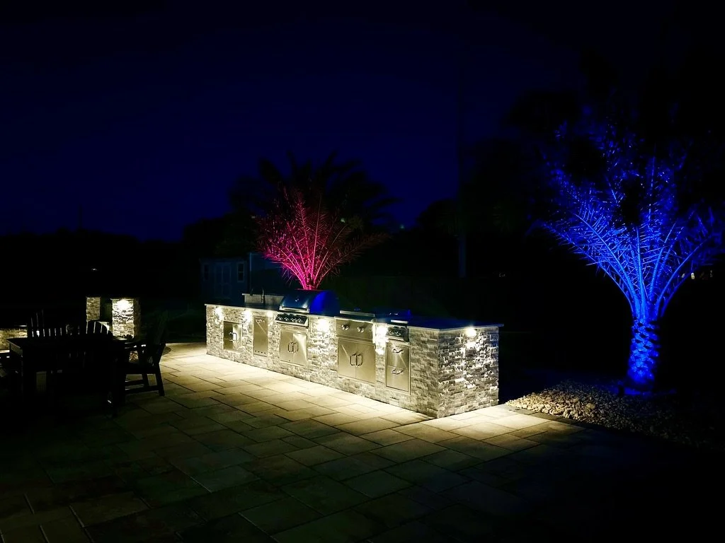 Outdoor kitchen with stainless steel appliances and stone countertop, illuminated at night with vibrant colored lighting on palm trees, creating a striking backyard ambiance.
