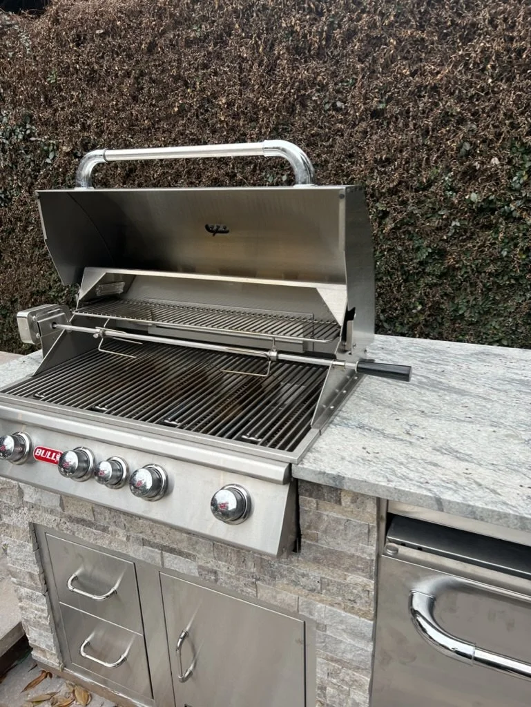 Close-up of a stainless steel outdoor grill with a stone countertop and storage drawers, set against a textured foliage background.