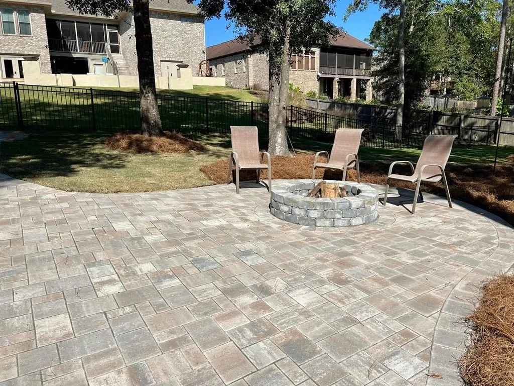 Backyard patio area featuring a stone fire pit surrounded by outdoor chairs, with a spacious lawn, trees, and nearby houses in the background.