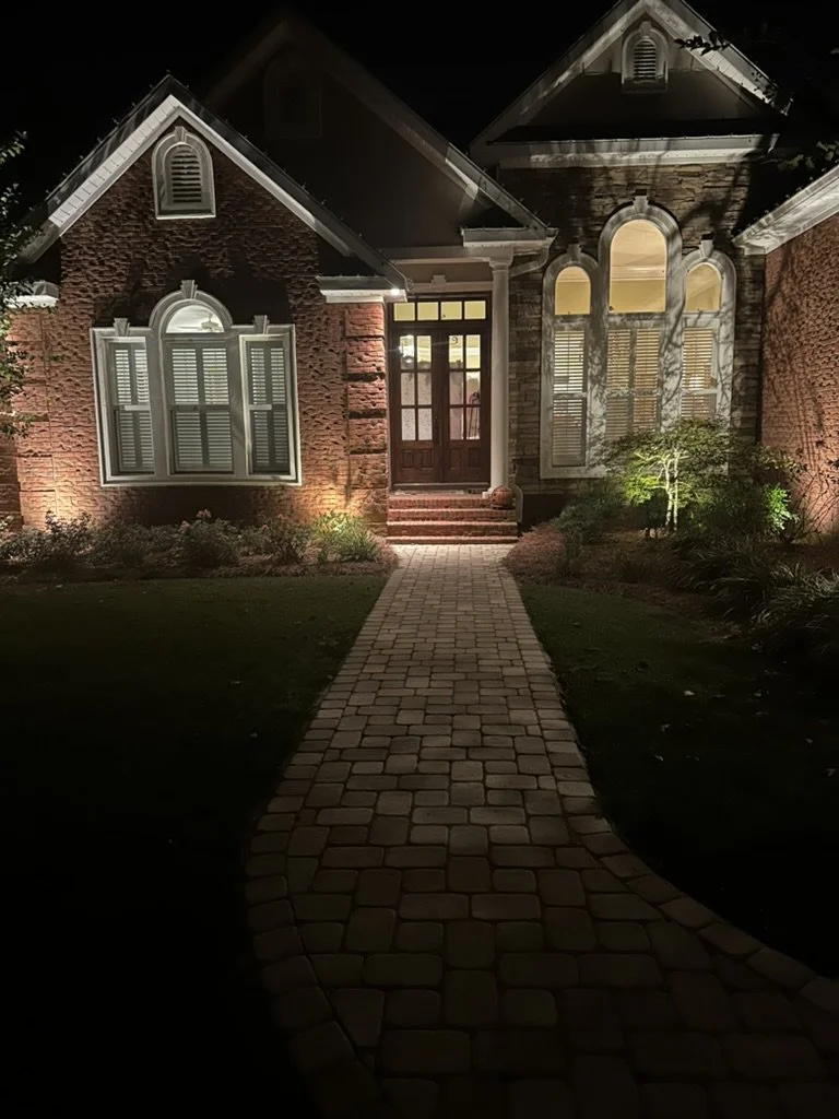Front entrance of a brick house with a well-lit stone walkway and landscape lighting highlighting the facade and garden at night.