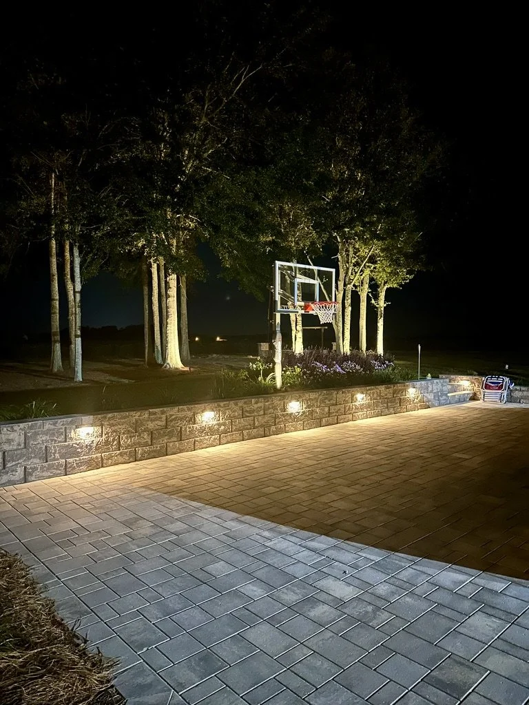 Outdoor basketball court at night with ambient lighting along stone wall and surrounding trees