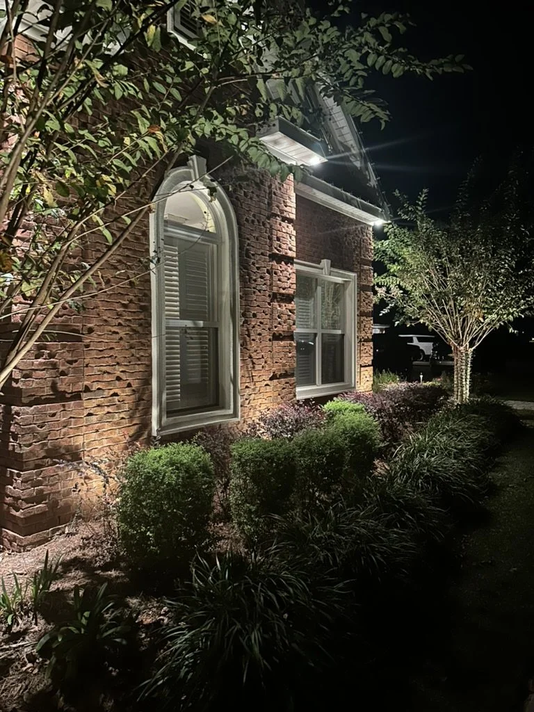 Close-up of a brick house exterior at night with landscape lighting highlighting arched windows, shrubs, and trees.