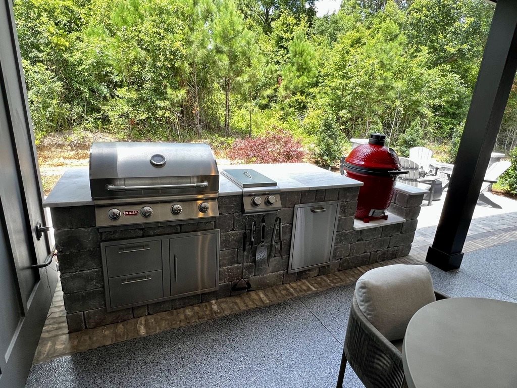 Outdoor kitchen featuring a stainless steel grill, a red ceramic smoker, and stone countertop, set against a lush green backdrop with outdoor seating